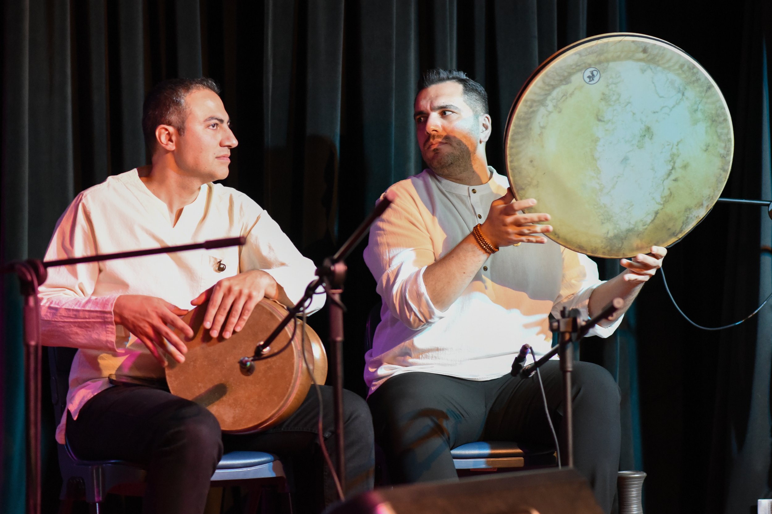 Two musicians playing traditional frame drums on stage, seated, with microphones in front of them.