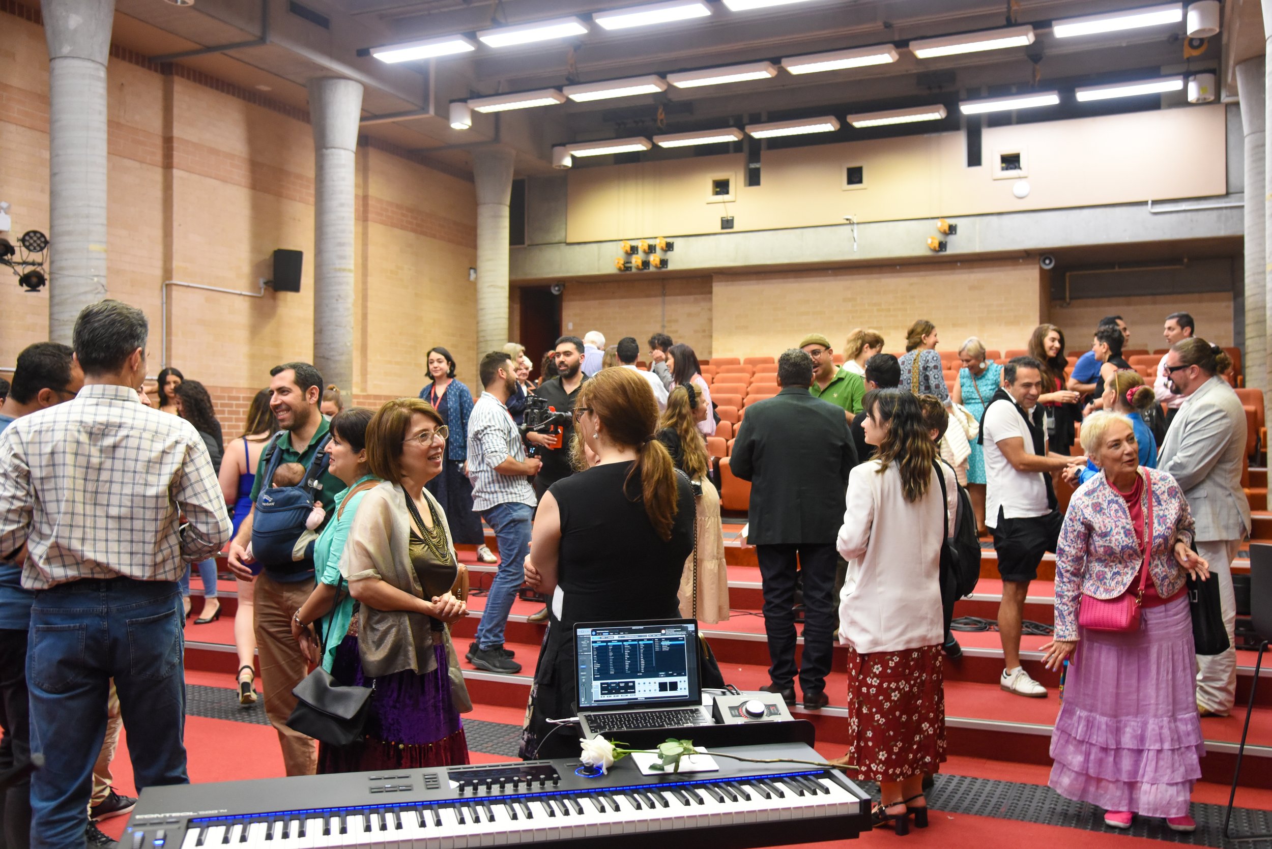 A group of people socializing in a large auditorium with red tiered seating and a digital keyboard and sound equipment in the foreground.