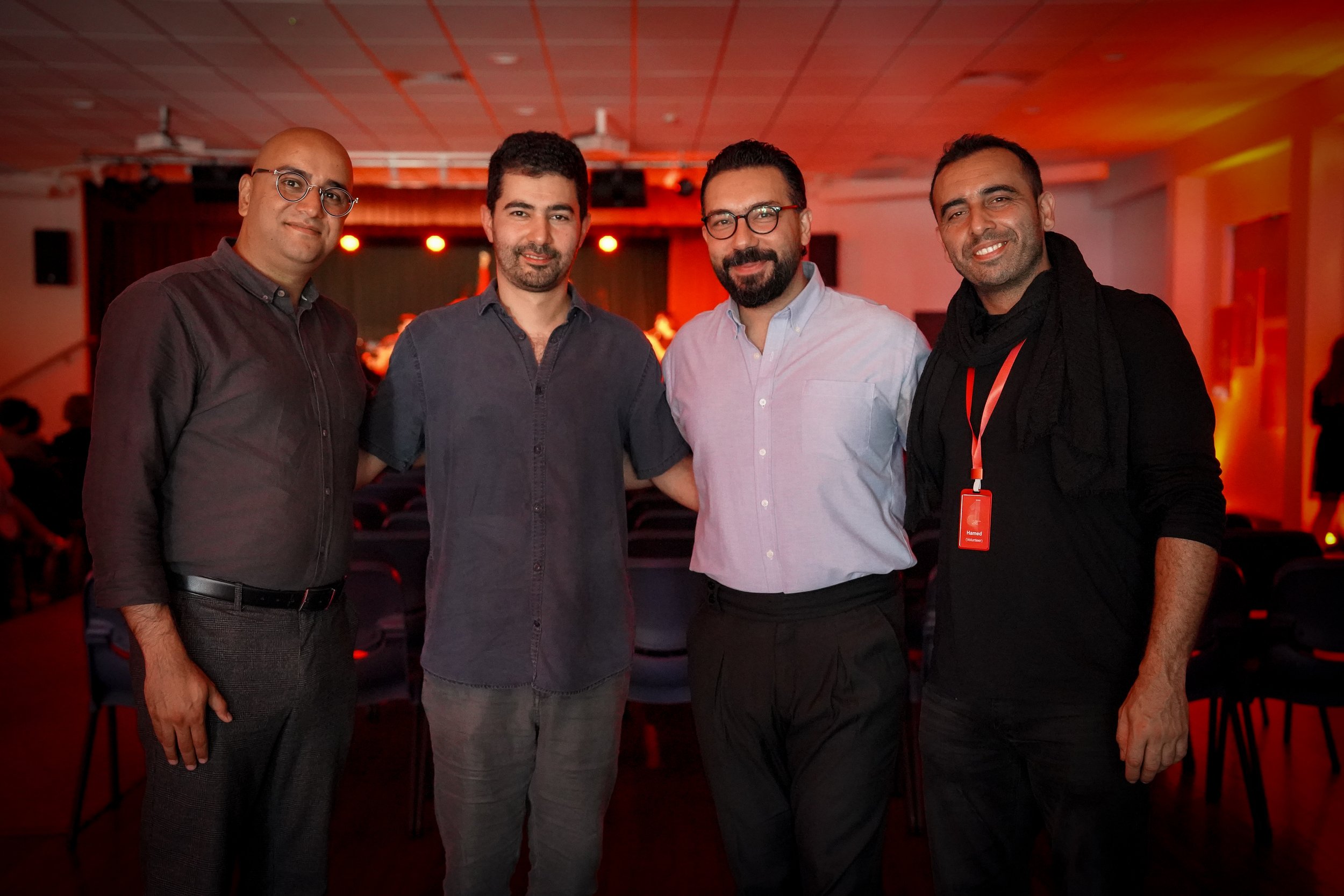 Four men standing together in a dimly lit room with red lighting, smiling at the camera. They are wearing casual and semi-formal attire. Chairs are visible in the background.