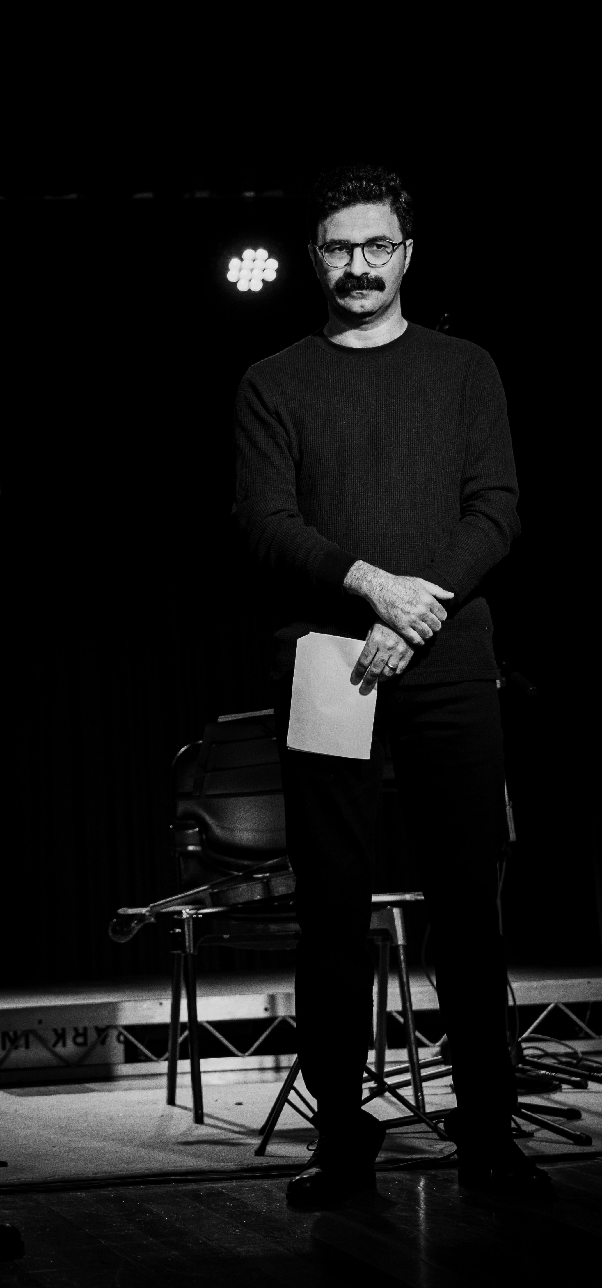 Black and white photo of a man standing on stage with a mustache and glasses, holding a paper in front of him, with a chair and microphone stand visible in the background.