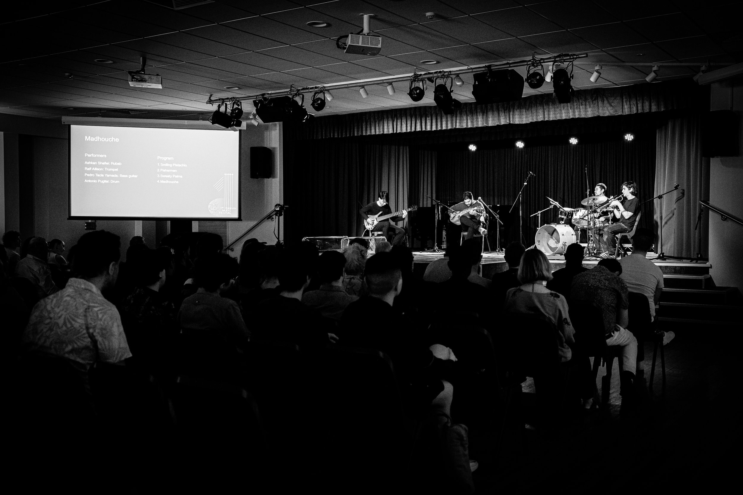 Black and white photo of a live music performance in a theater with audience seated. Four musicians are on stage, playing instruments including a guitar and drums. A projector screen is displaying information above the performers.