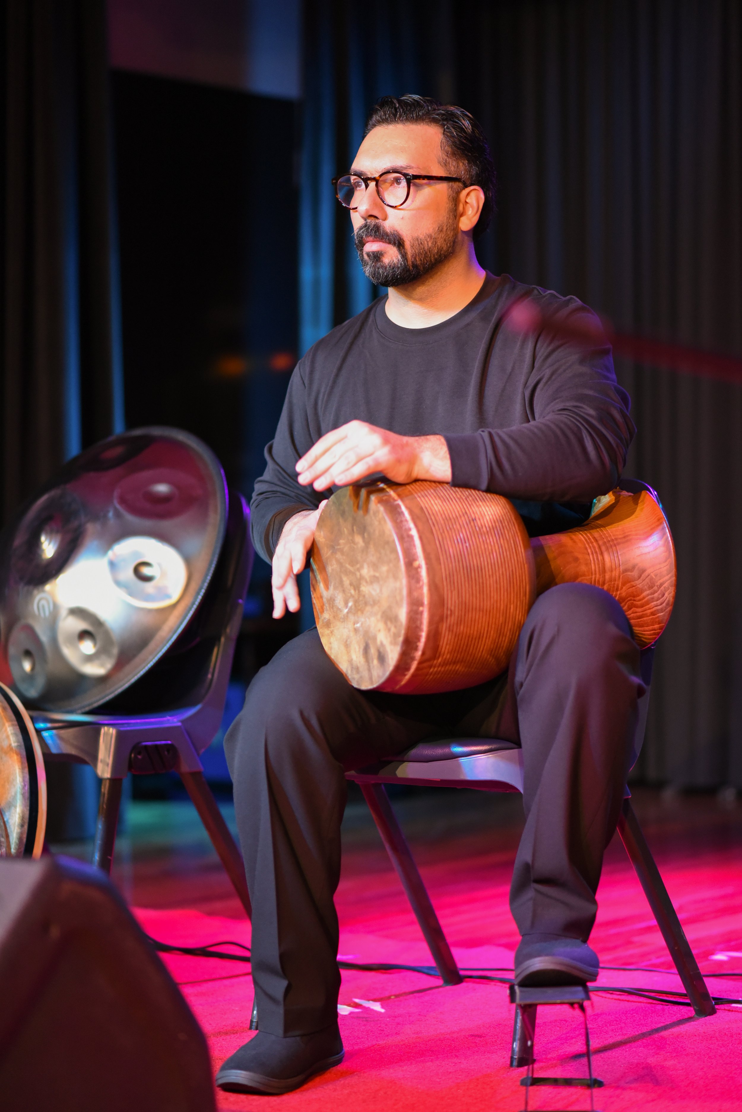 A musician seated on a chair playing a traditional drum on stage, with a handpan instrument visible beside him.
