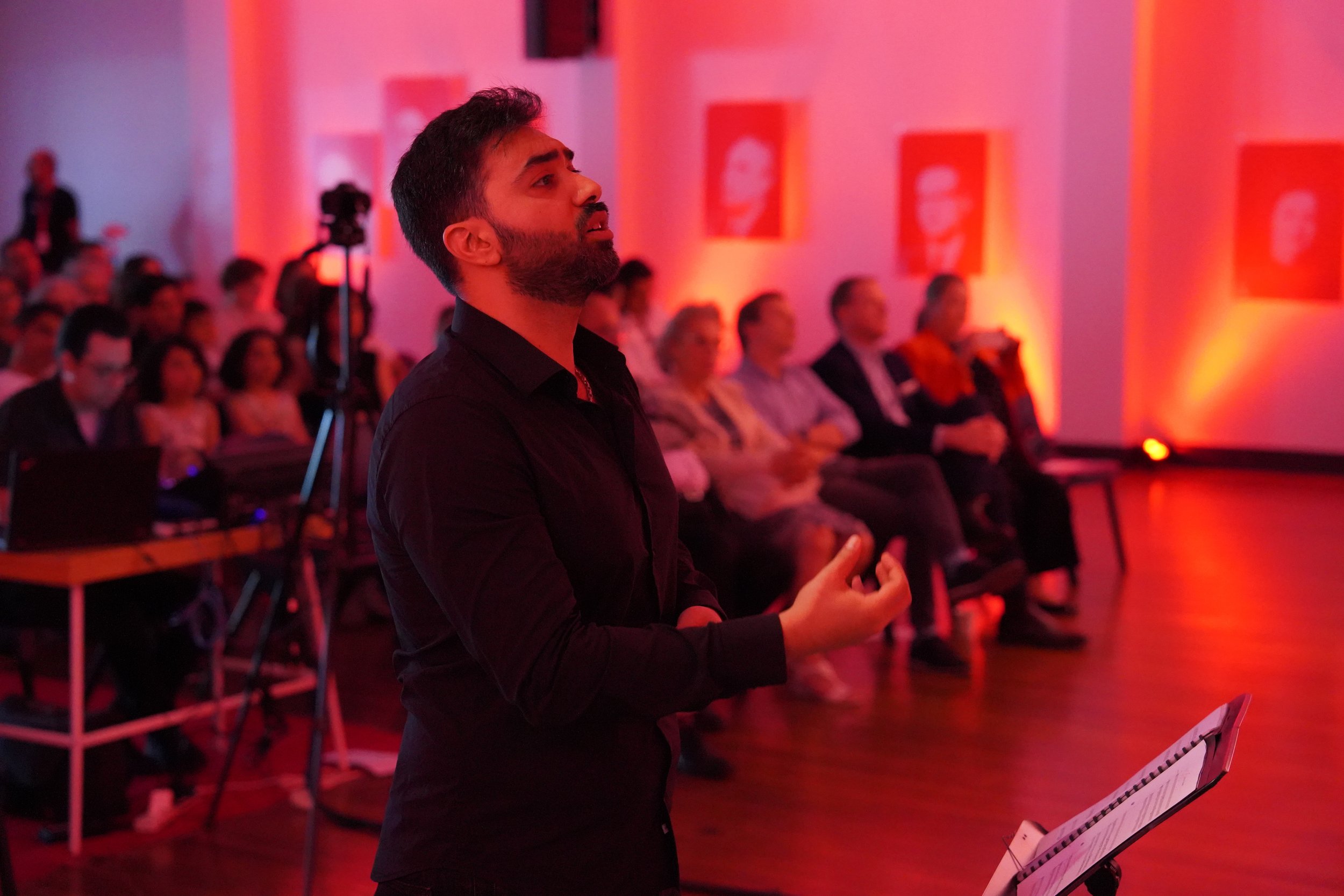 Person speaking at an event, red ambient lighting, audience seated, camera and equipment visible.