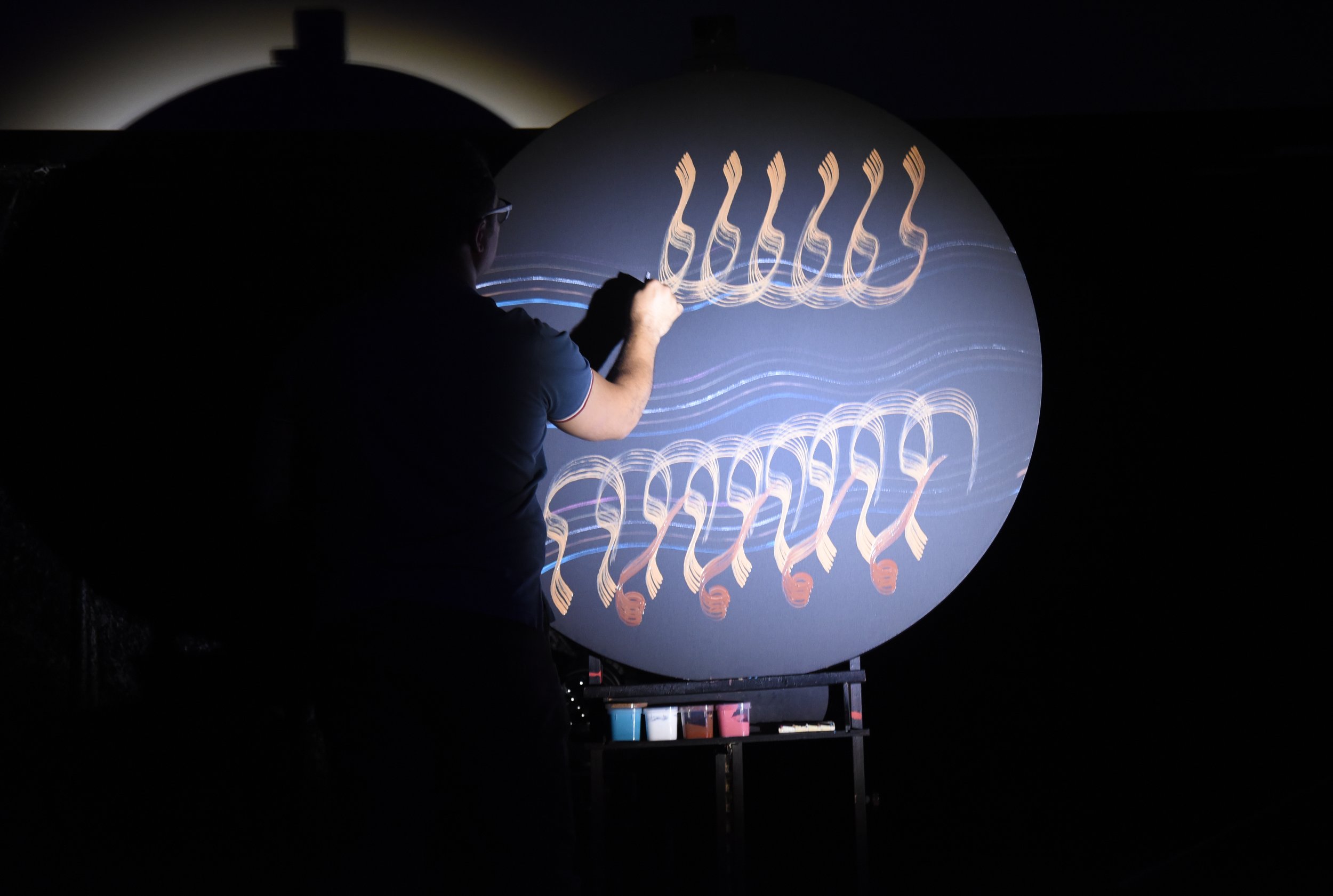 Person painting on a large circular canvas in dim lighting, creating abstract orange and blue patterns.