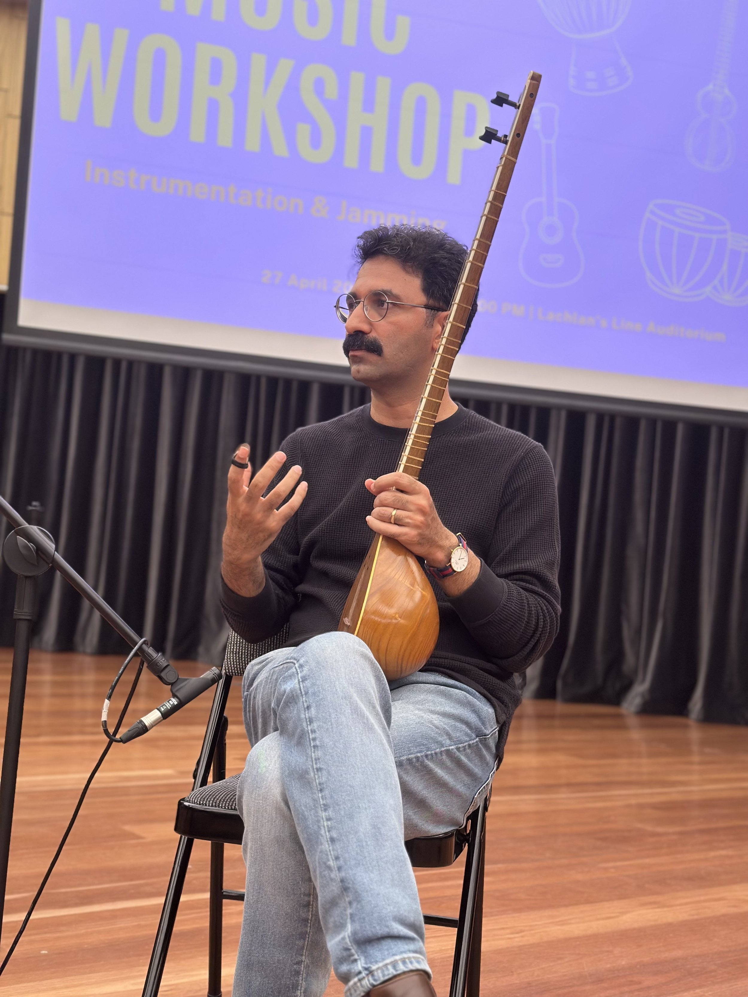 A man seated on a chair holding a stringed musical instrument, discussing at a music workshop. There's a microphone in front of him and a projector screen with the text 'Music Workshop - Instrumentation & Jamming' in the background.