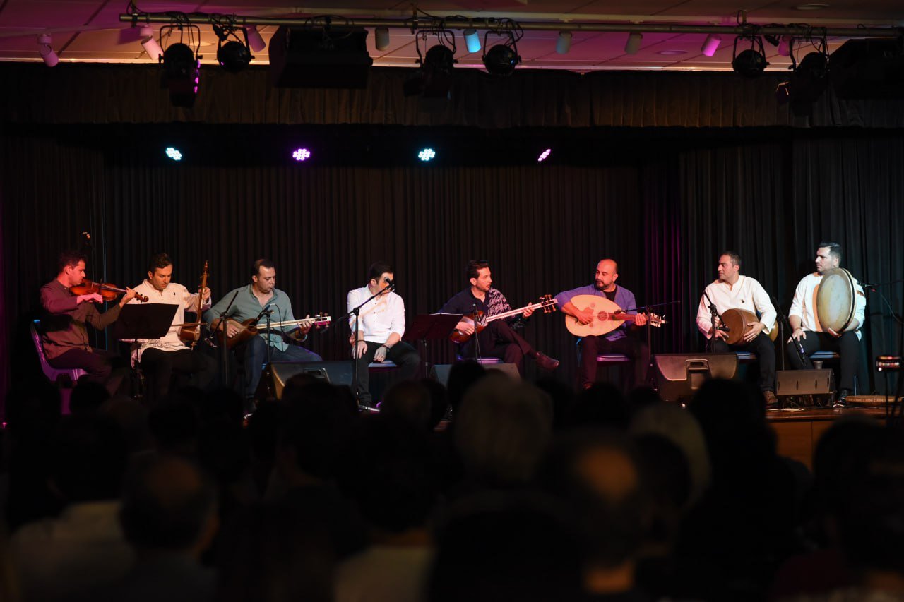 Musicians performing on stage with various string and percussion instruments in front of an audience.