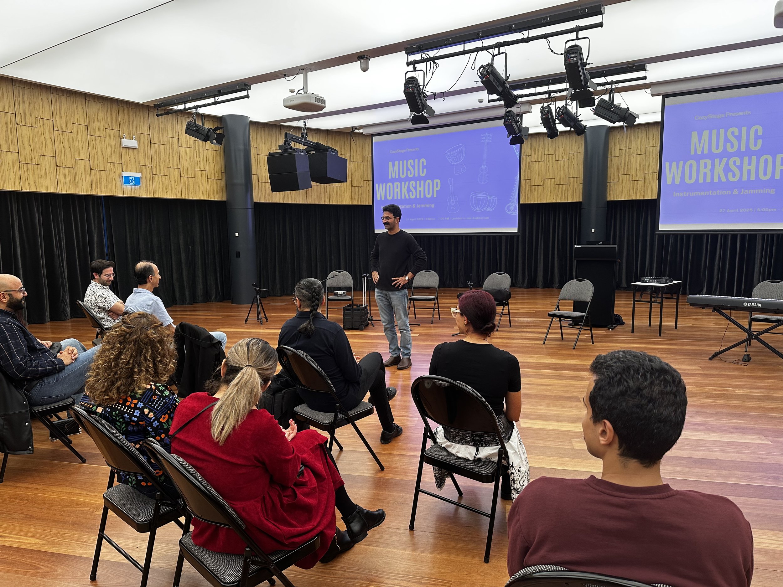 A person leading a music workshop in a room with wood flooring. Several people are seated facing the front, with two large screens displaying 'Music Workshop' and musical instruments on the wall. Chairs and a keyboard are visible in the background.