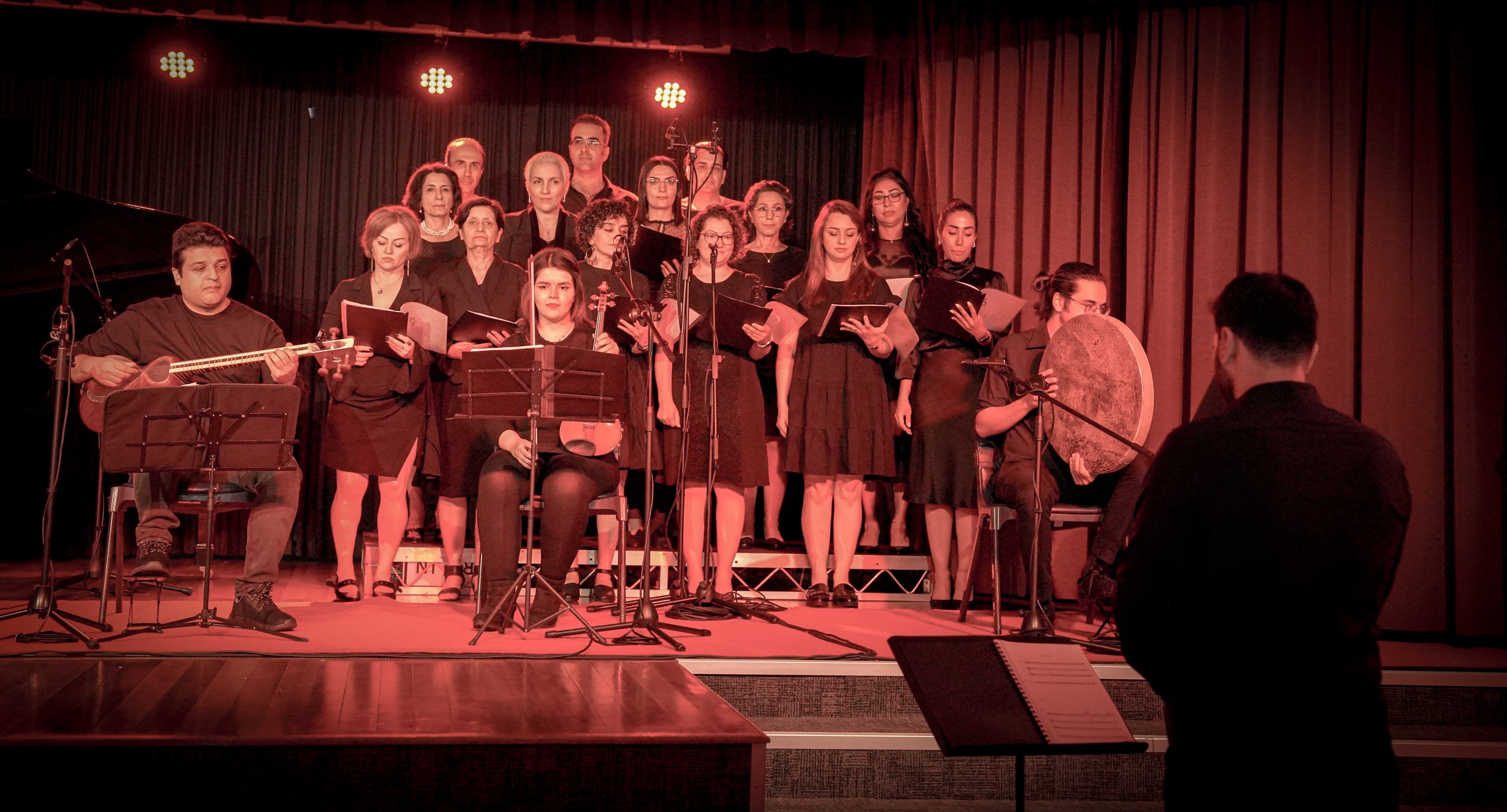 A choir on stage with a conductor in front, a guitarist, a violinist, and a percussionist playing a frame drum. The choir members hold open books and appear to be mid-performance. The lighting is red, creating a warm ambiance.