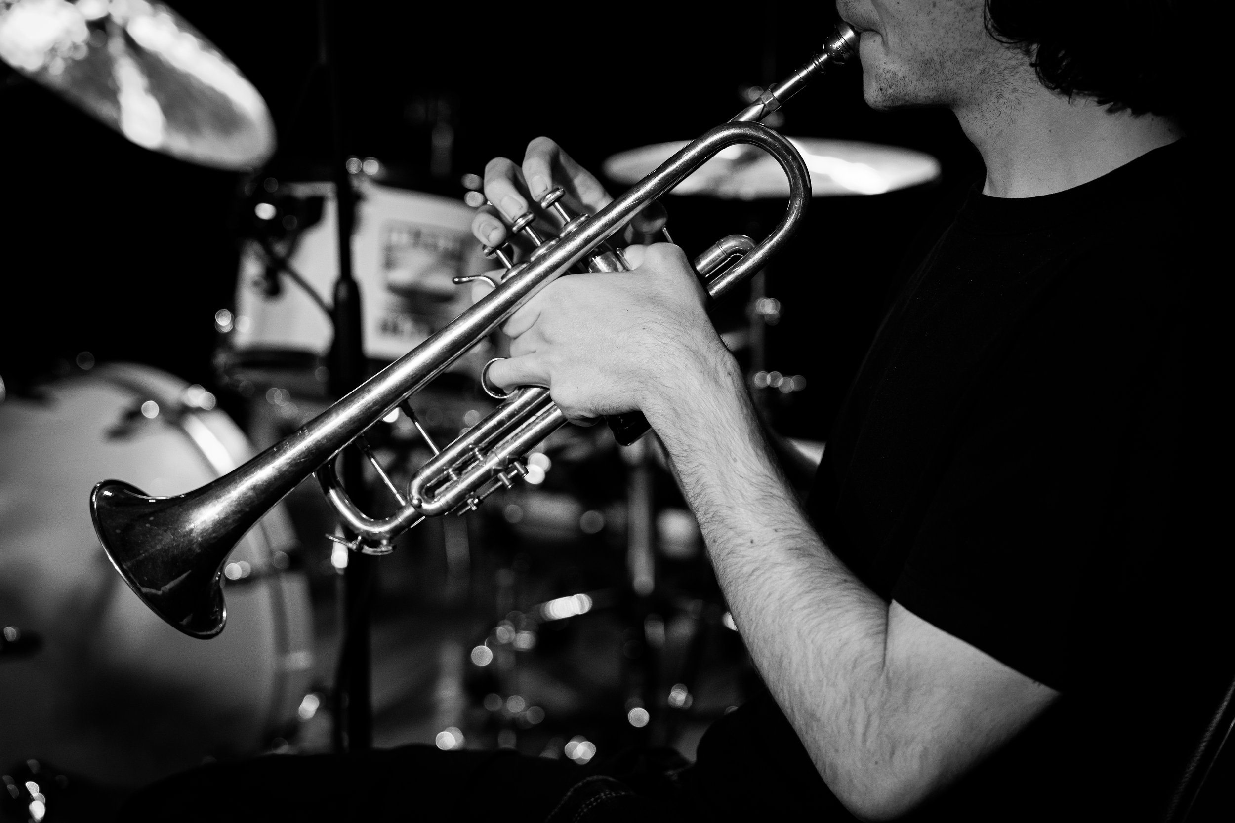 Black and white photo of a person playing a trumpet with a drum set in the background.