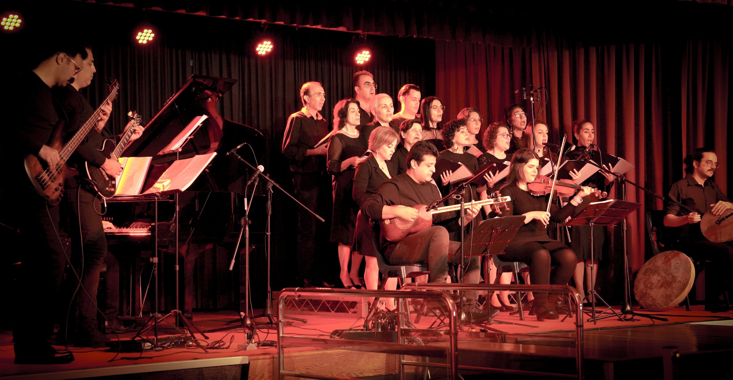 A choir and musicians performing on stage with a bass guitarist, violinist, and percussionist playing under red stage lights.