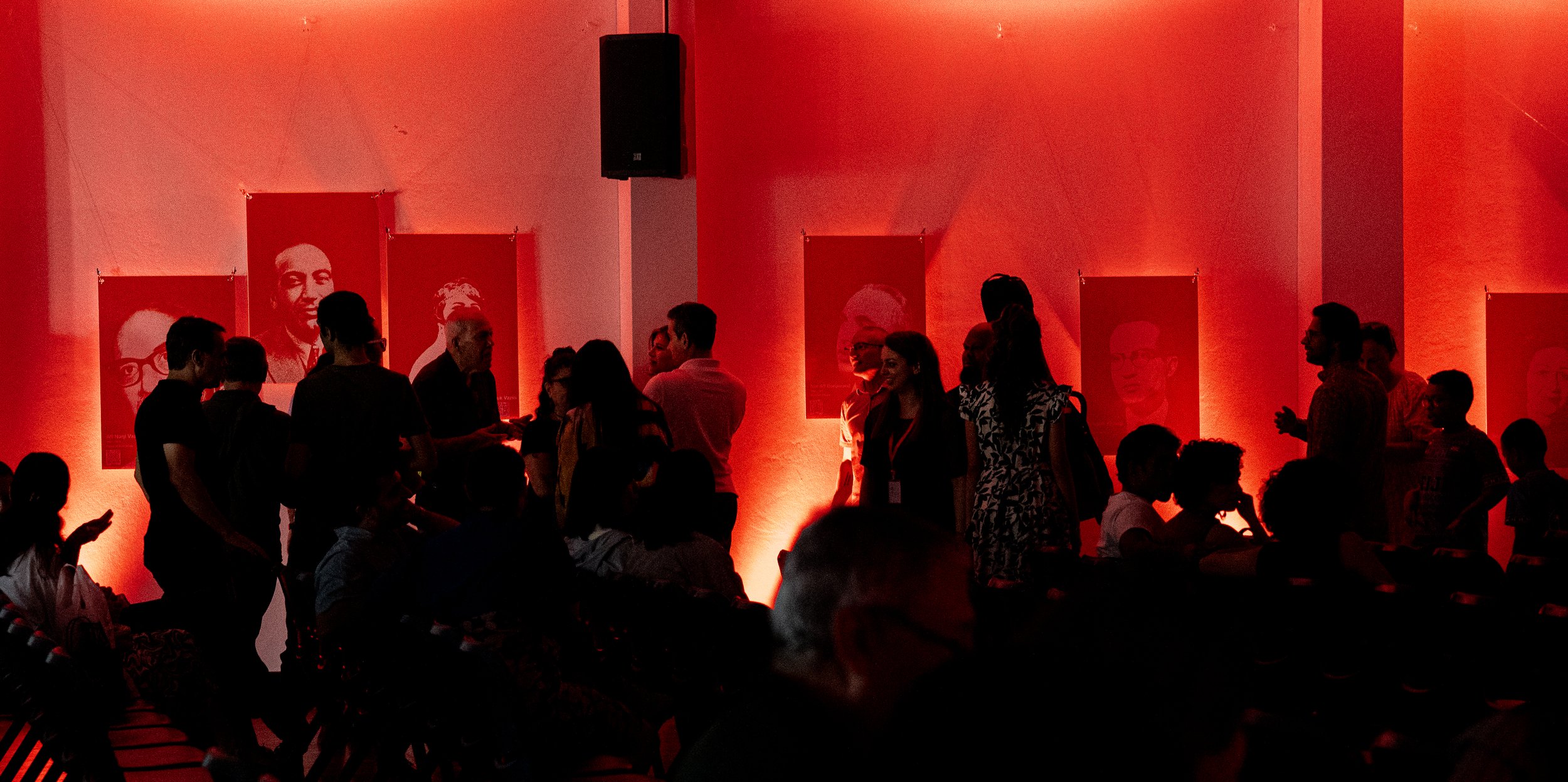 Dimly lit room with red lighting, people gathered and conversing, wall adorned with large portraits, possibly of historical or cultural figures.