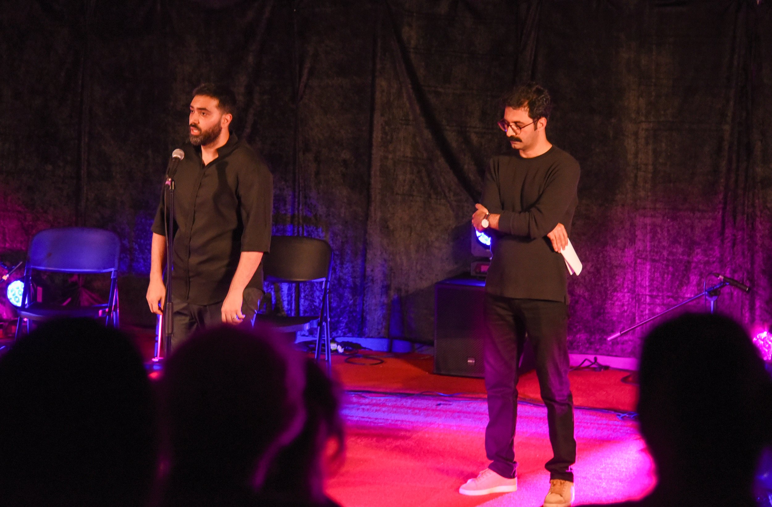 Two men performing on stage with microphones, audience in the foreground, colorful lighting.