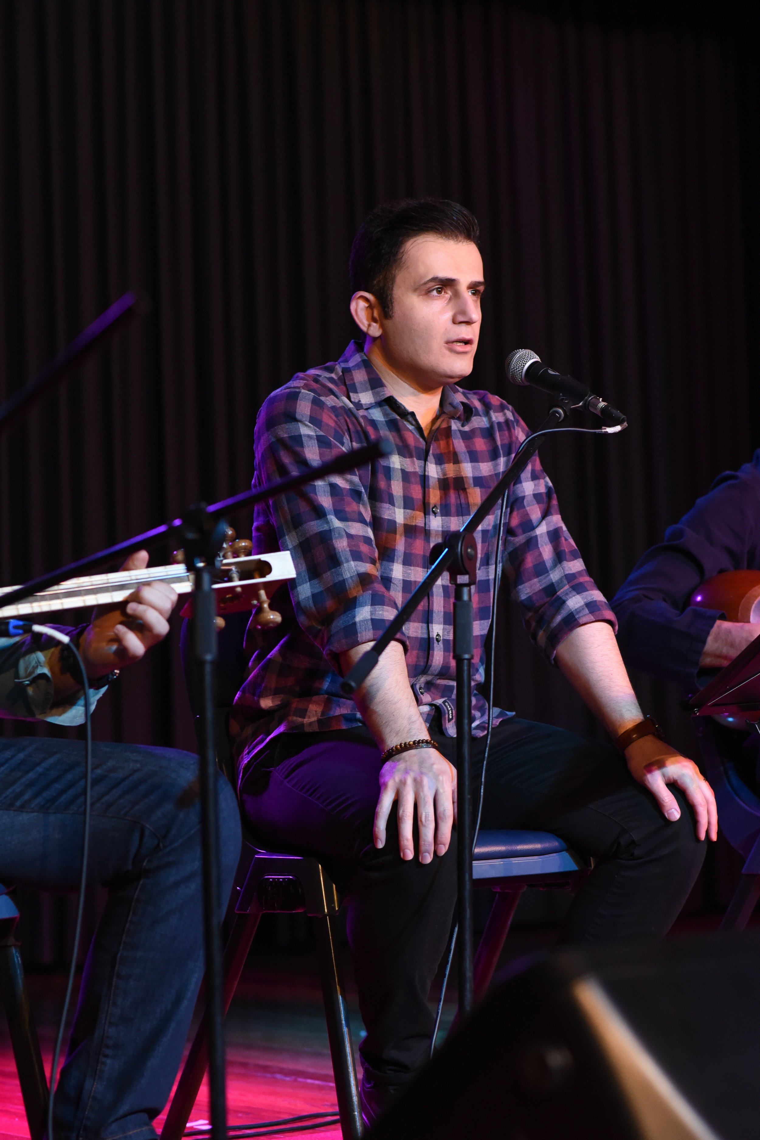 Man in checkered shirt sitting on stage with a microphone, playing music.