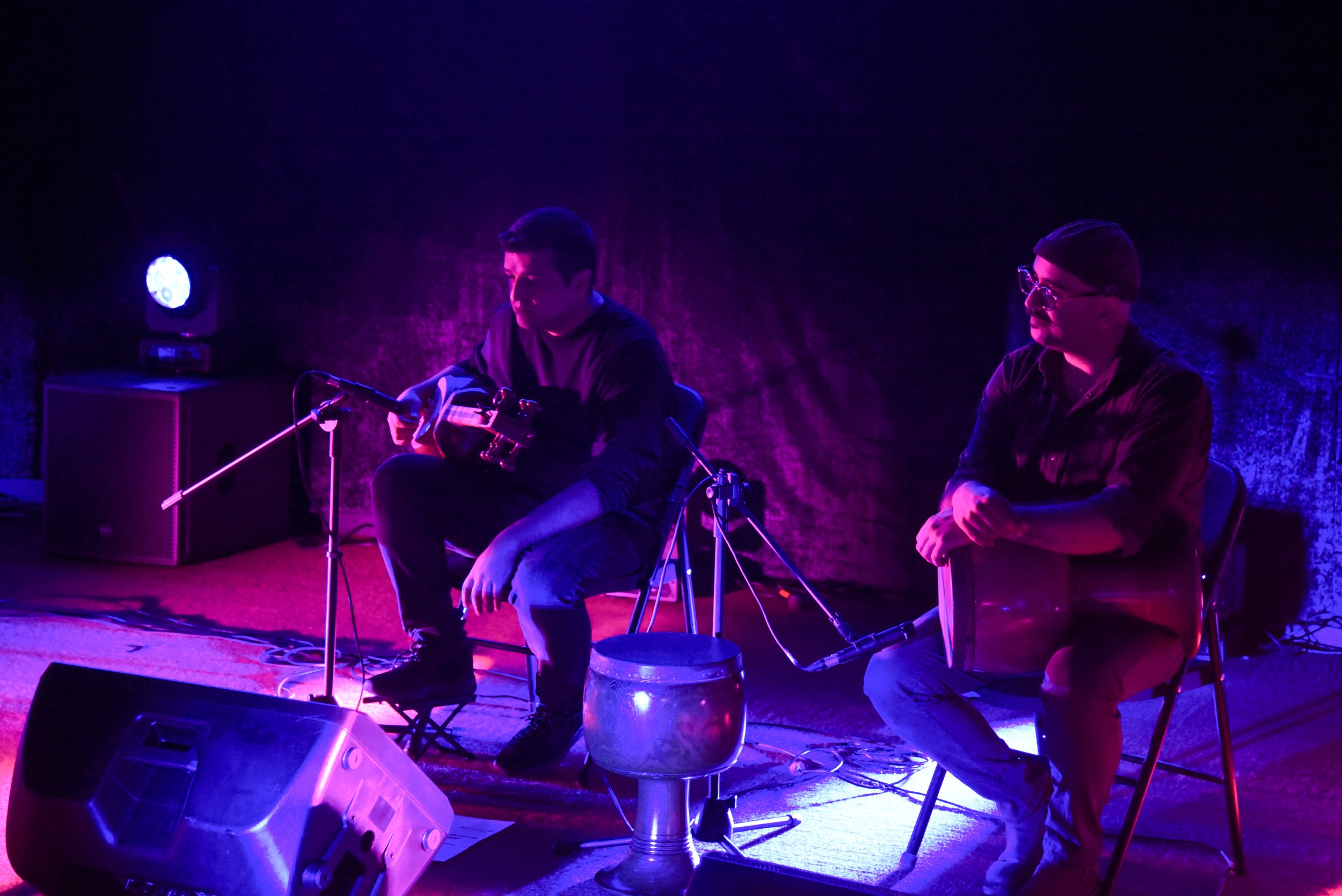 Two musicians performing on stage with purple lighting, one playing a string instrument and the other with a drum. Microphones and audio equipment are visible.