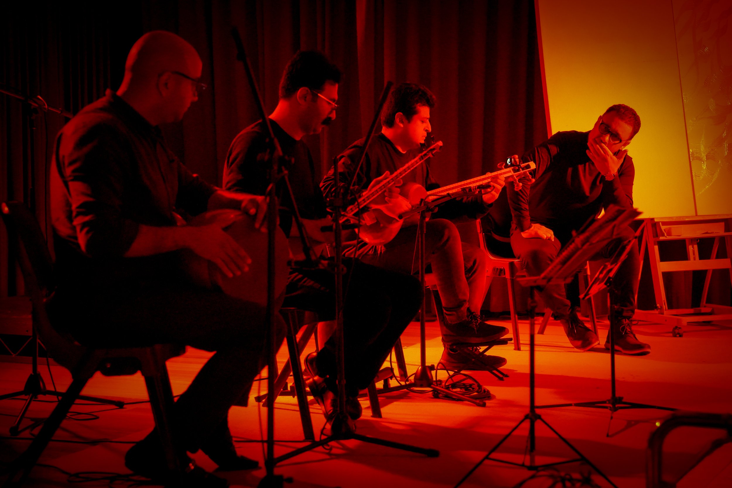 Four musicians performing on stage with Iranian instruments under red lighting.