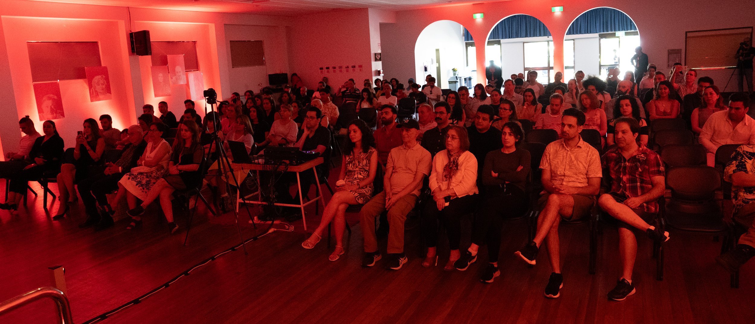 Audience attending an indoor event with red ambient lighting, featuring seated people and a projector setup.