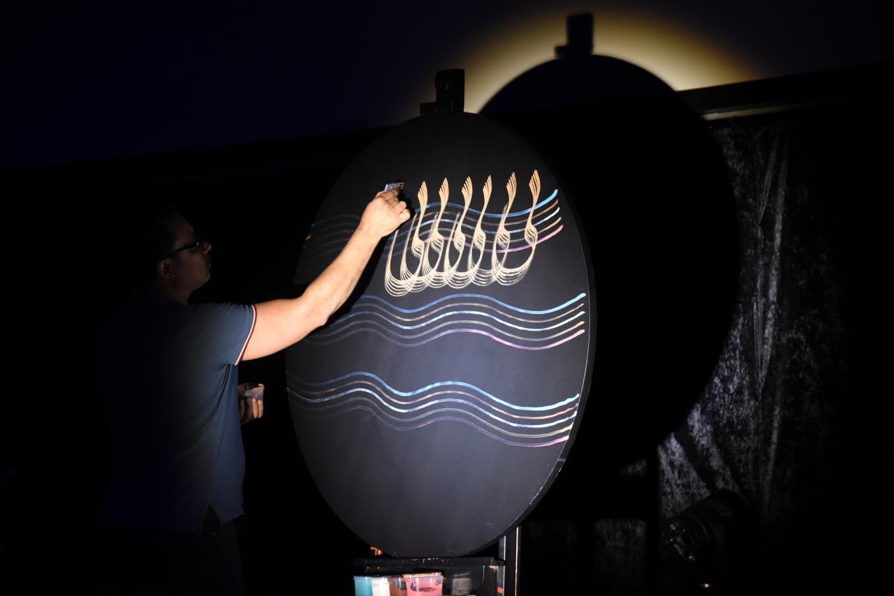 A person painting calligraphy on a round, dark board with flowing wave patterns and decorative script. The scene is dimly lit.