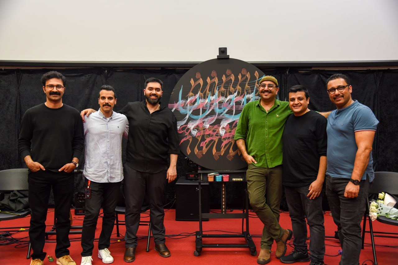 Group of six men posing in front of an abstract painting on a large circular canvas.