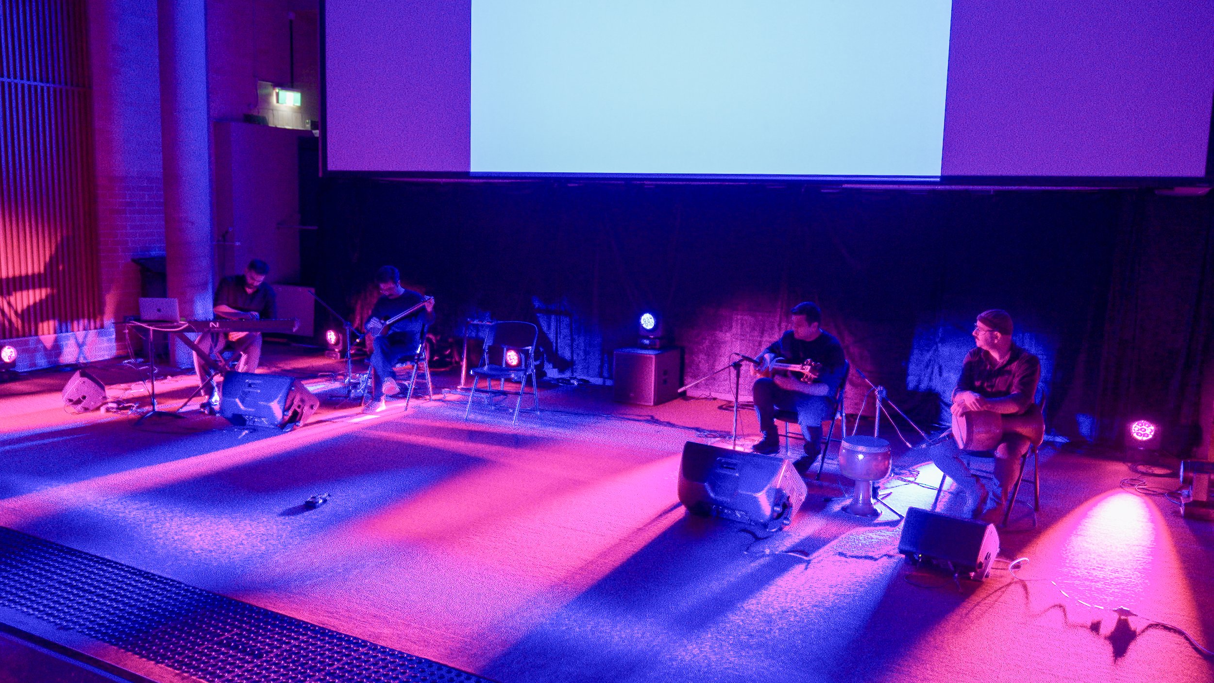 Musicians performing on stage with colorful lighting, sitting on chairs, playing instruments like a keyboard and guitar.