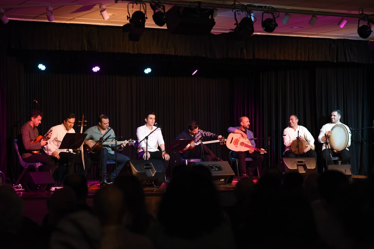 Group of musicians performing on stage with various string and percussion instruments, under soft stage lighting.