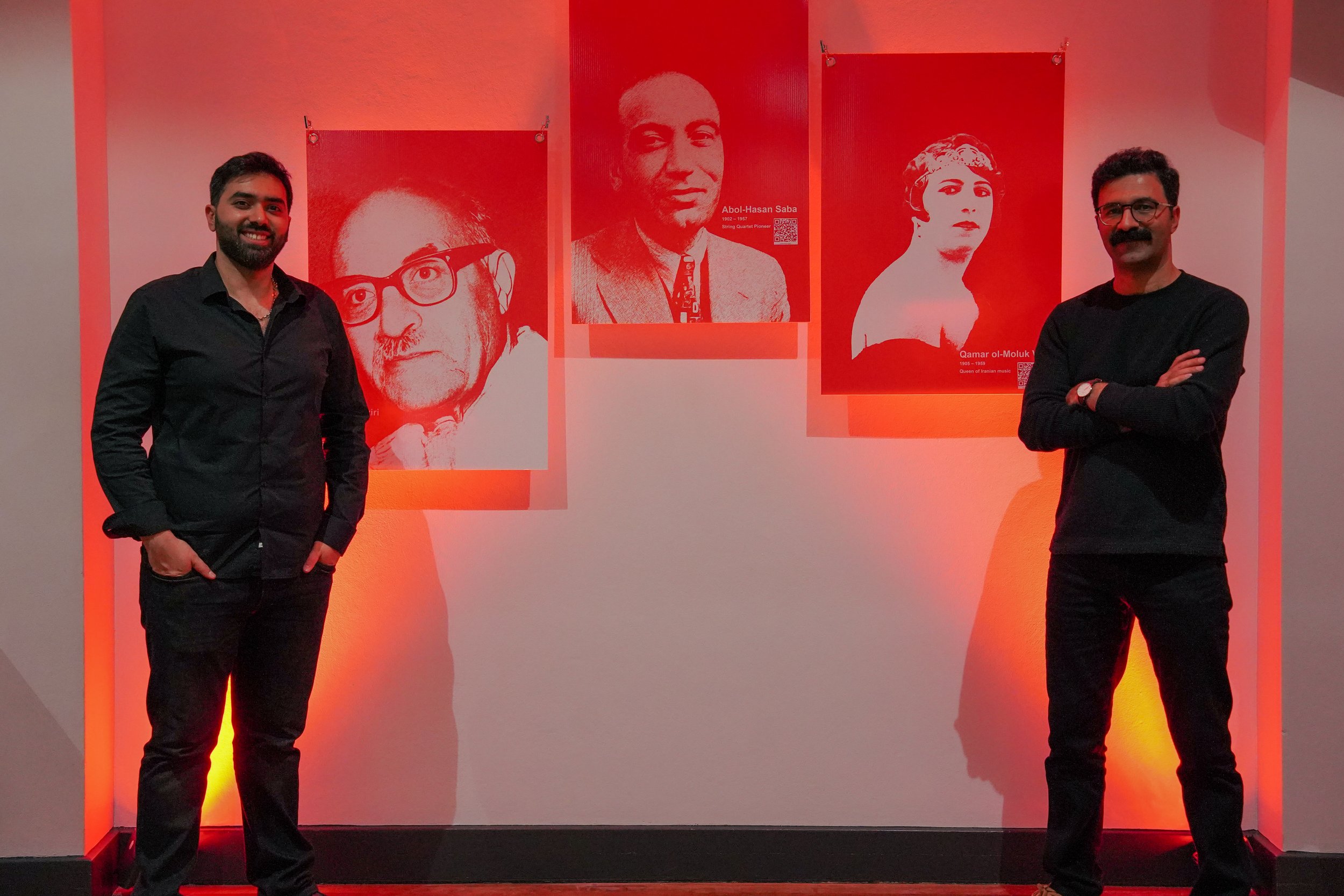 Two men stand on either side of three red-toned posters featuring black and white portraits of historical figures, illuminated by red lighting in a gallery setting.