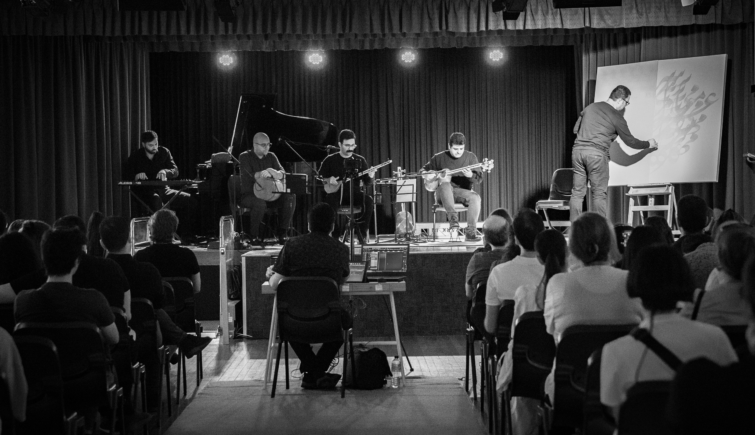 Black and white photo of a musical performance on stage with musicians playing instruments and an artist drawing on a canvas, viewed by an audience.