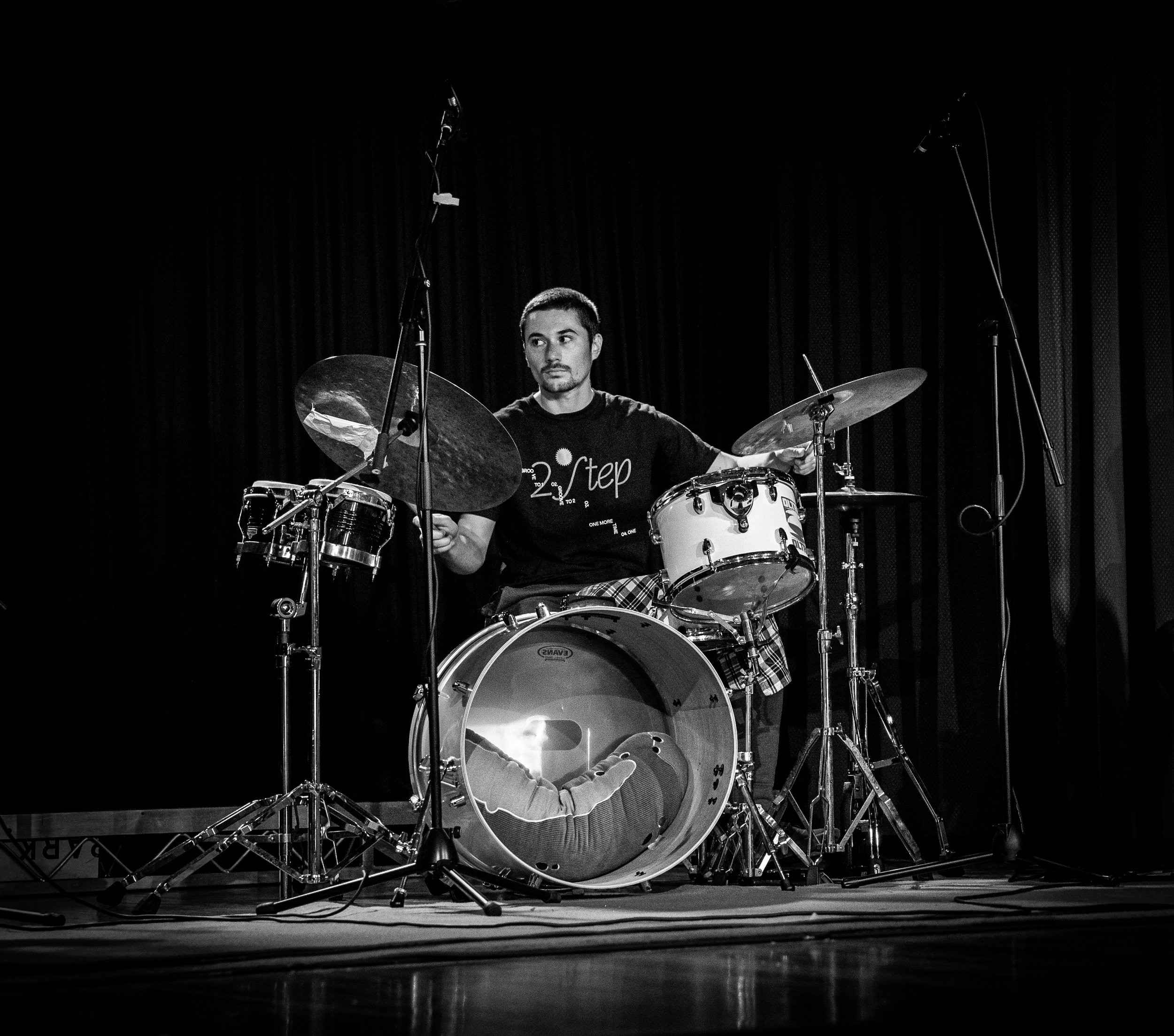 Black and white photo of a drummer playing a drum set on stage with a microphone in the foreground.