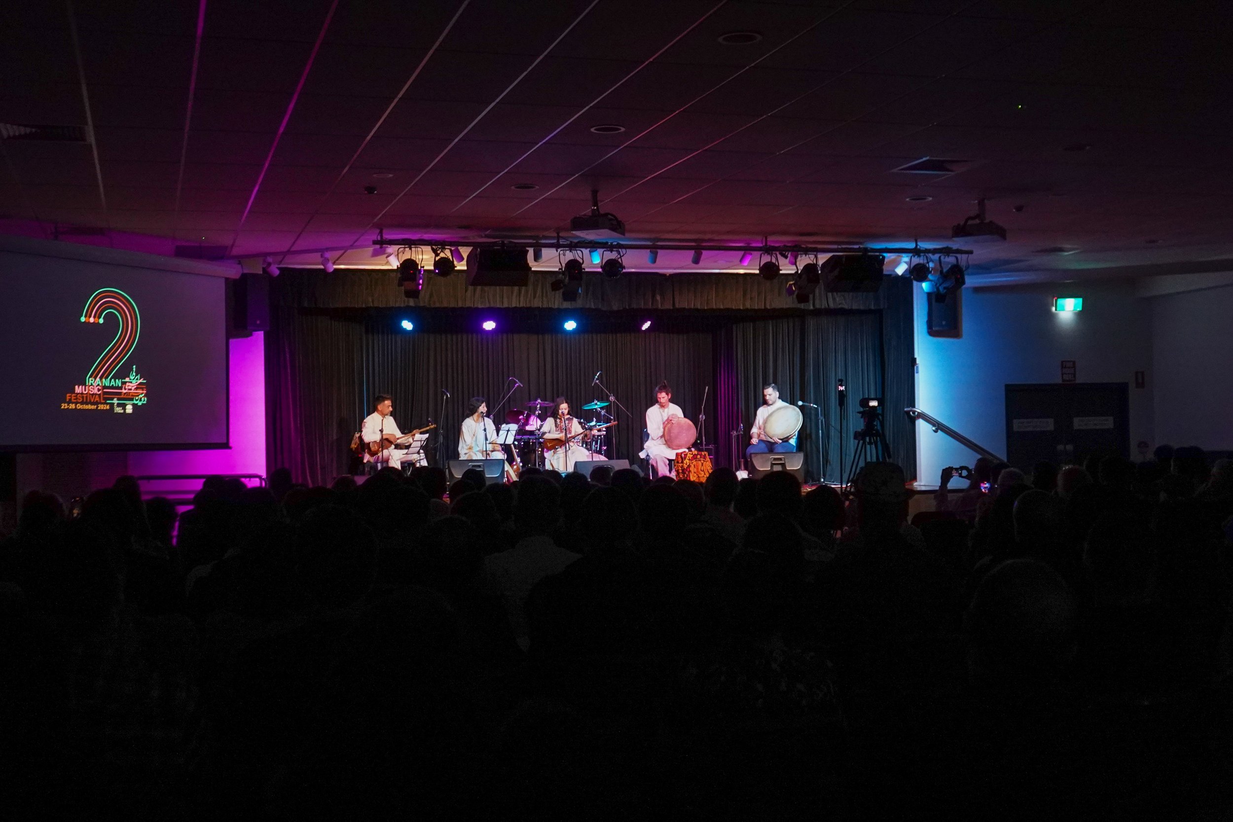 Musicians performing on stage at the Iranian Music Festival 2018 with colorful stage lighting, audience in silhouette, and a screen displaying the festival logo and date.