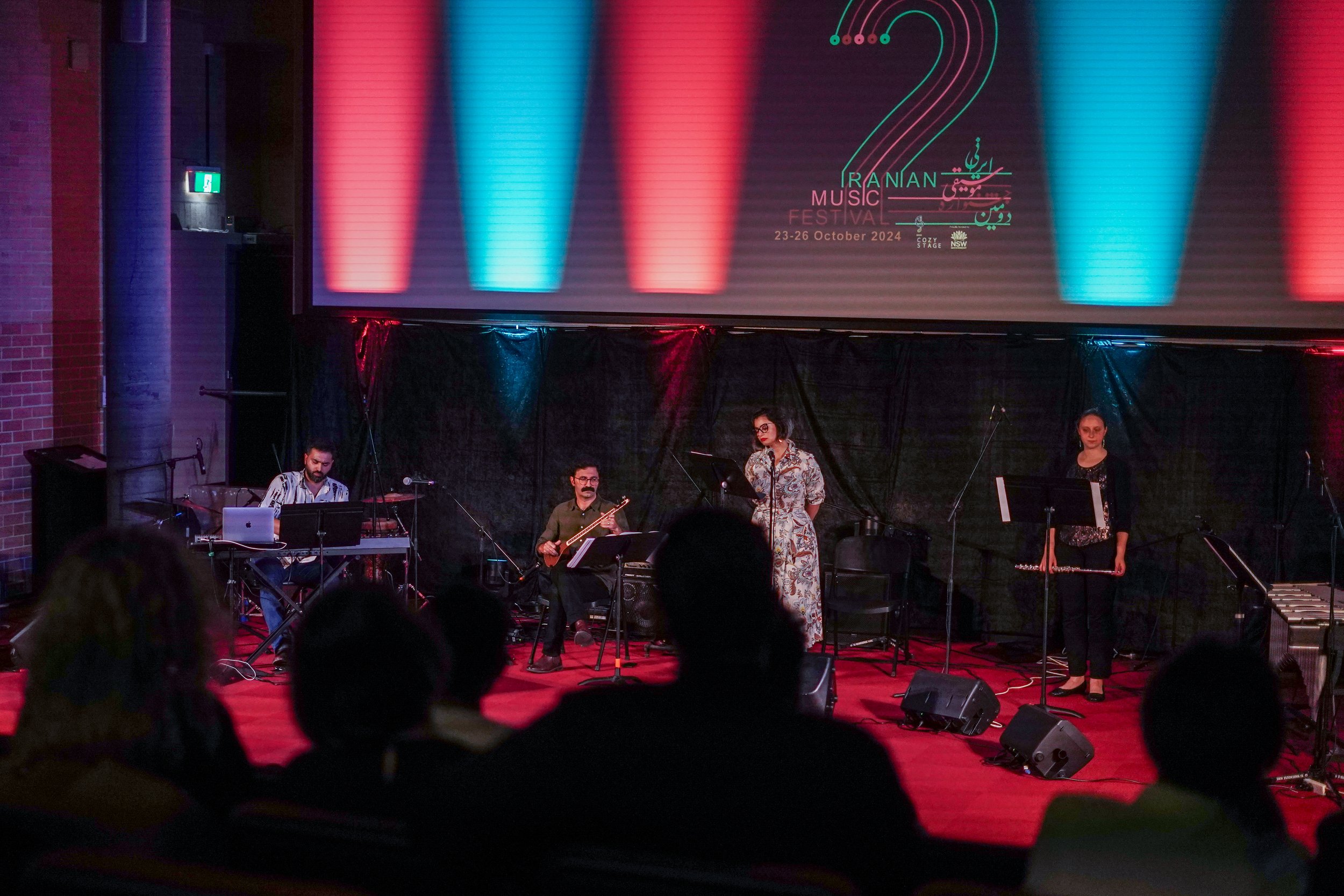 Performers on stage at the Iranian Music Festival with red and blue lighting and instruments like a keyboard and guitar. Event details on screen behind them, dated October 23-26, 2024.