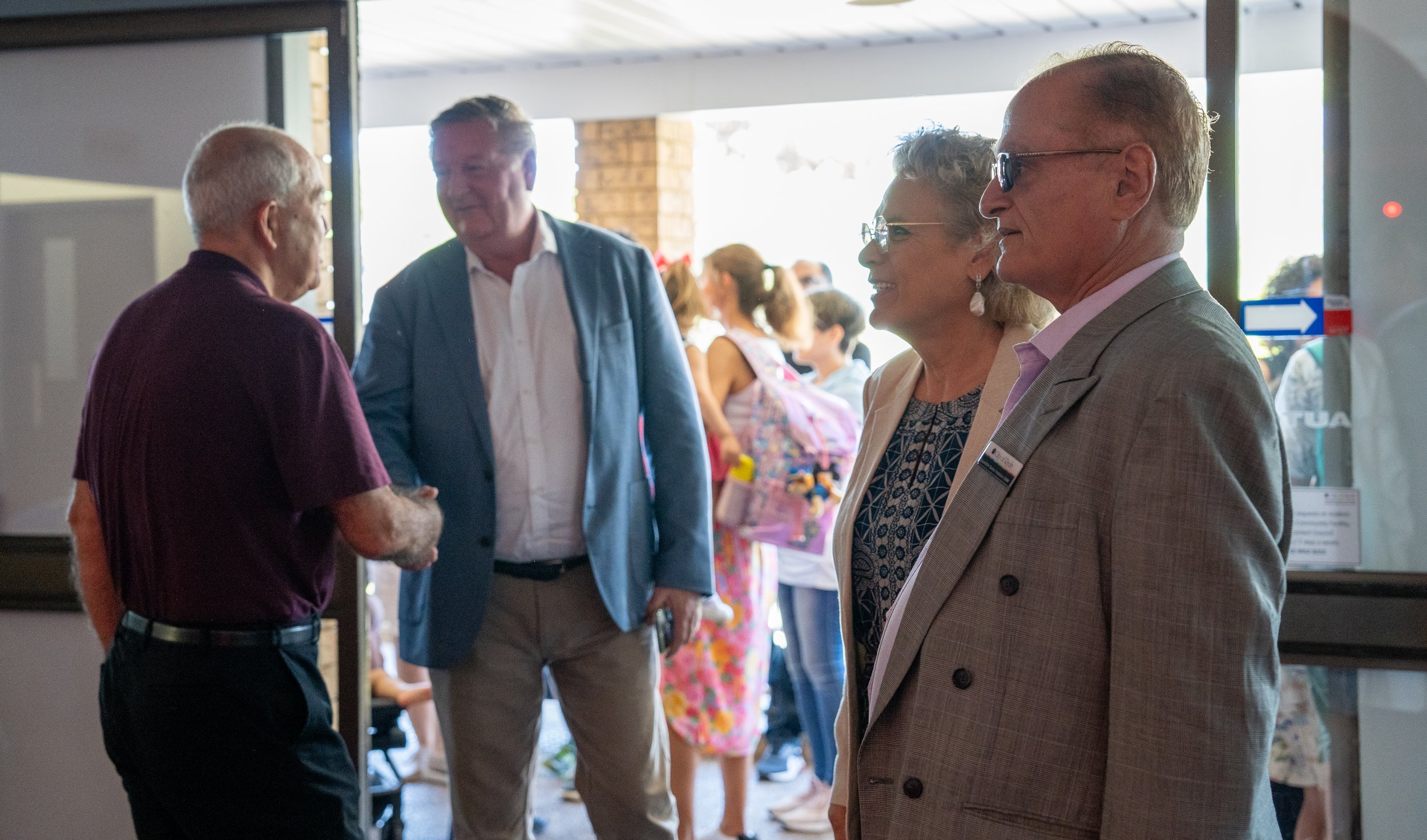 Group of people, including a man in a maroon shirt shaking hands with another in a blue blazer, near an entrance at an event.