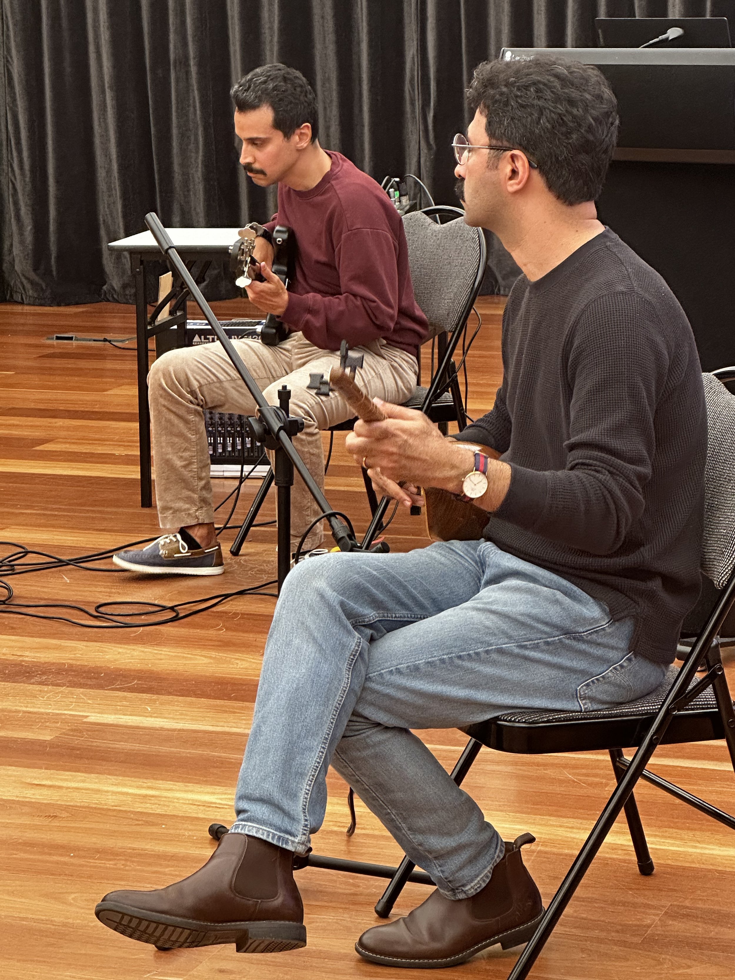 Two men sitting on chairs, playing musical instruments, possibly in a rehearsal room with wooden flooring and a black curtain background.