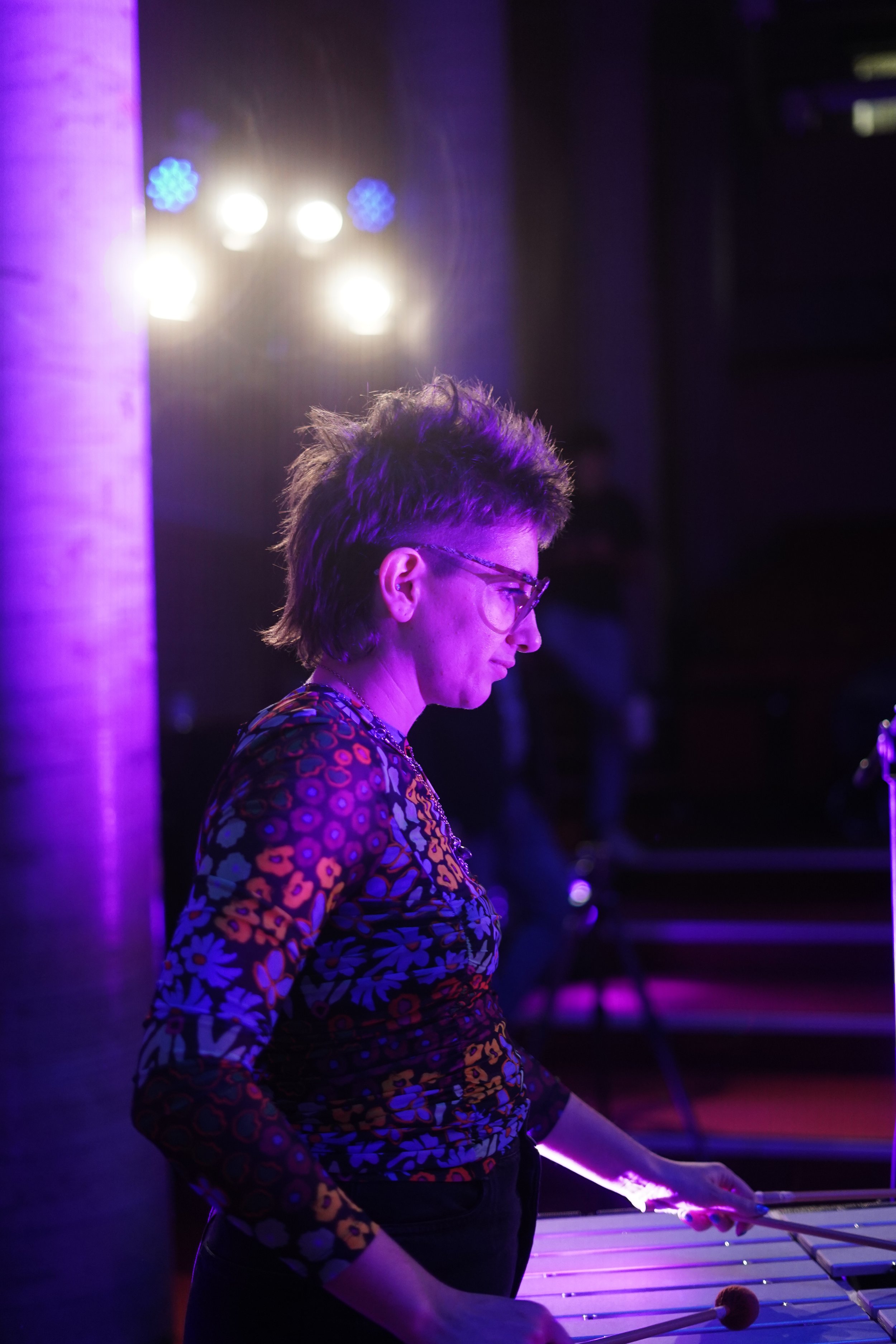 Musician playing xylophone under purple stage lights, wearing colorful floral shirt and glasses