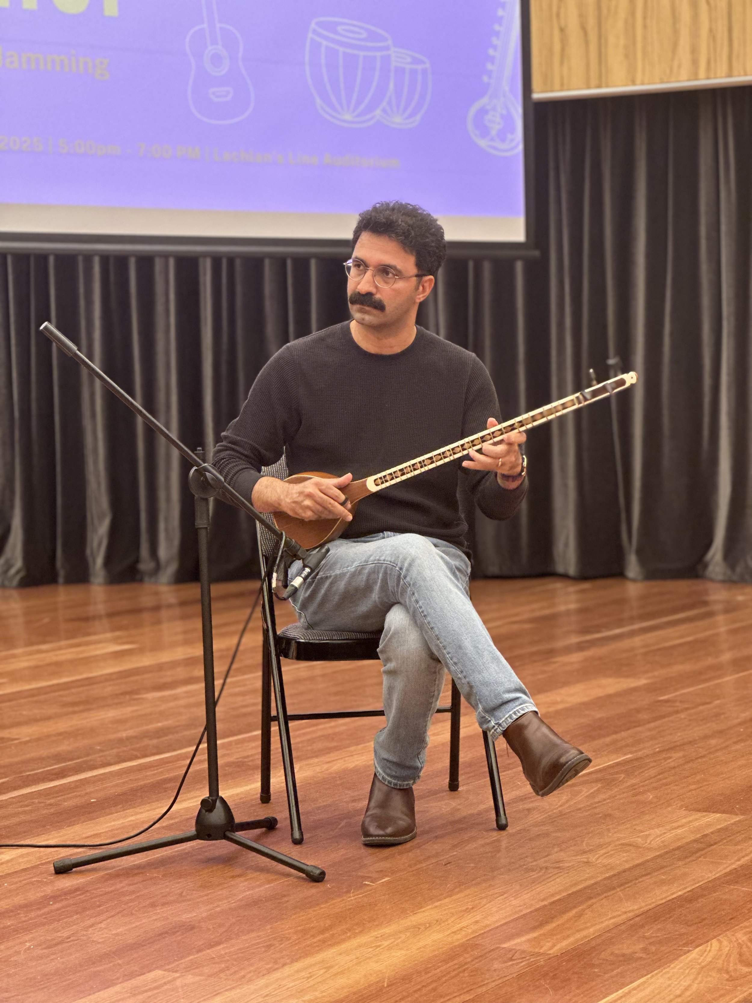 Man seated on a chair playing a tar instrument with a microphone in a performance setting; wearing glasses, black sweater, jeans, and brown boots. Background includes a projection screen with musical instruments' images.