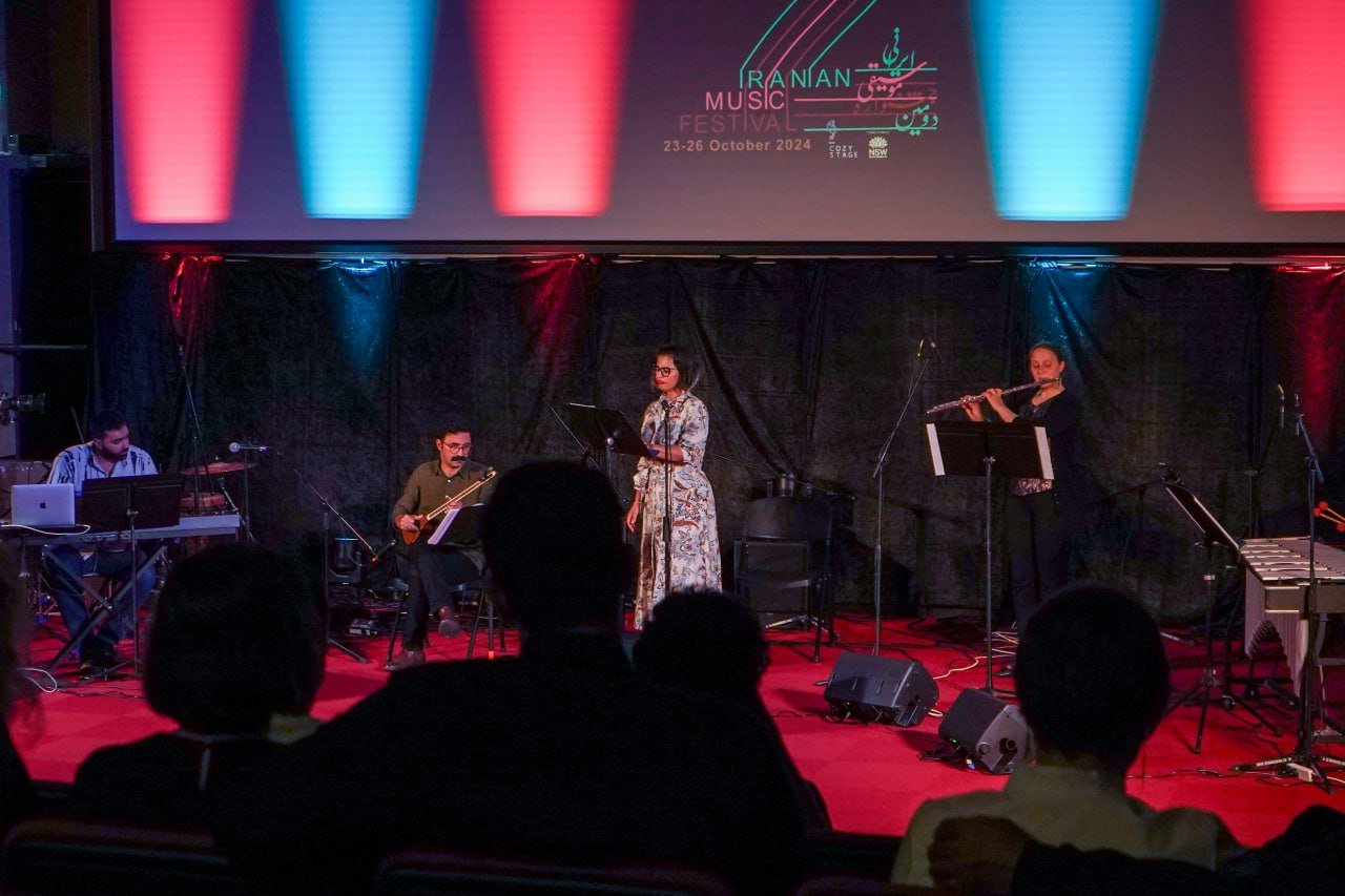 Musicians performing on stage at the Iranian Music Festival, with a backdrop showing festival details for October 2024. The stage is lit with red and blue lights, and the performers include a keyboardist, traditional string player, singer, and flutis