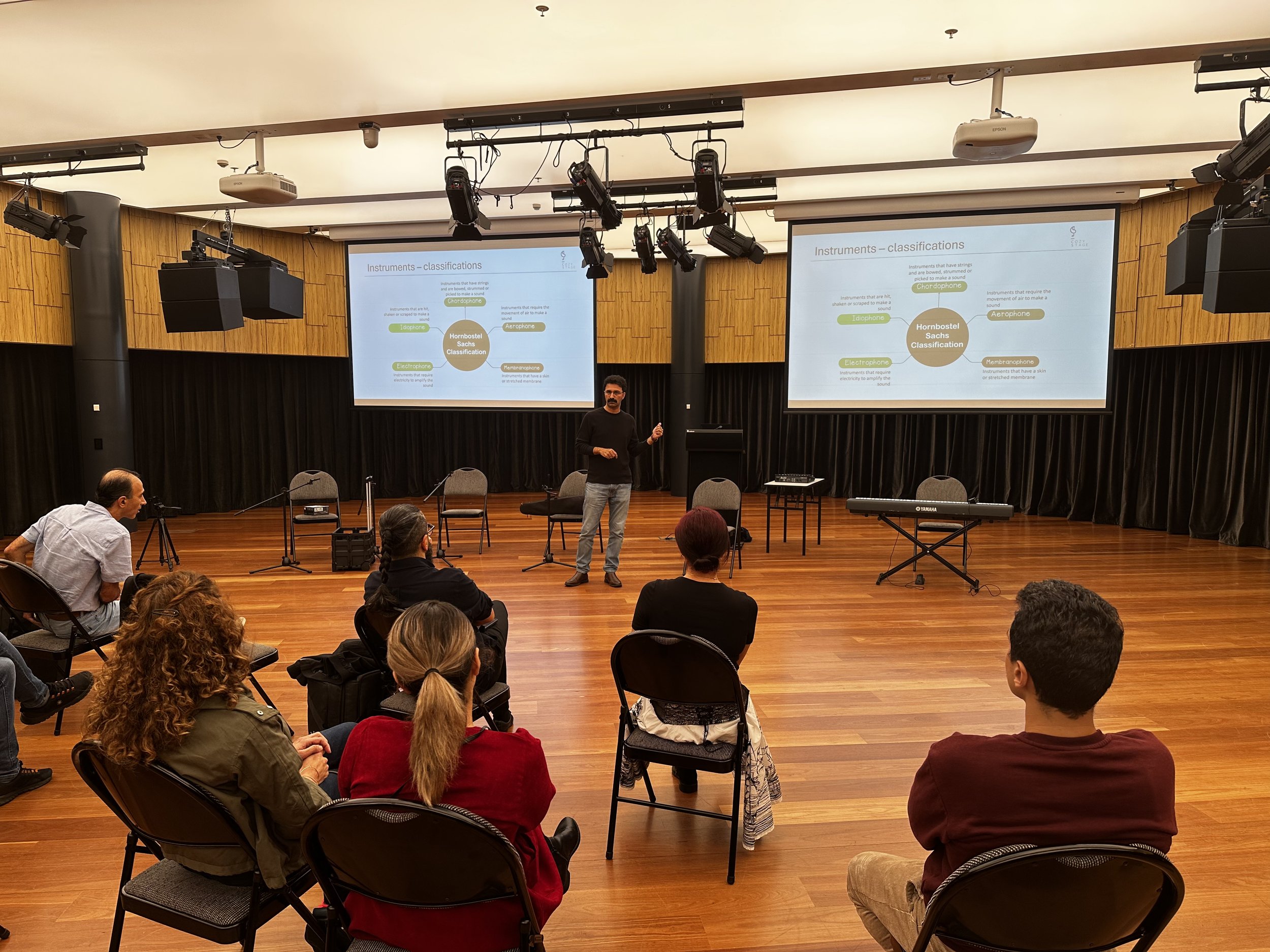 A person giving a presentation in a classroom or seminar room setting. The presenter stands in front of a screen displaying a presentation slide titled "Instruments - classifications," with various categories of instruments shown. Several people are