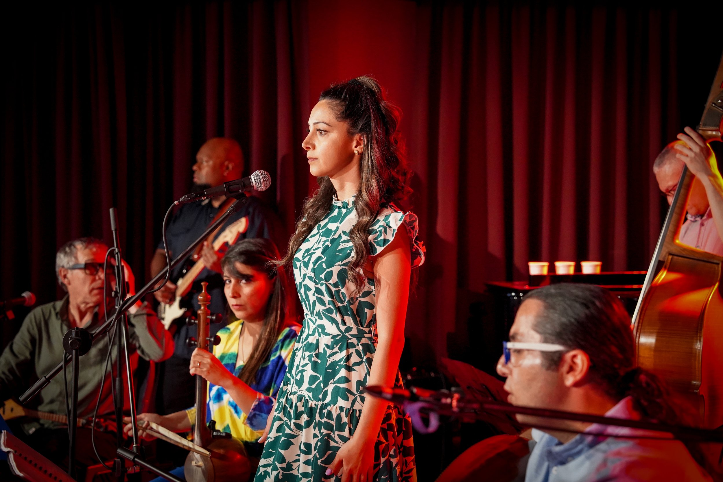 A band performing on stage with a female vocalist and various musicians playing instruments such as a bass guitar and string instruments, under red lighting.