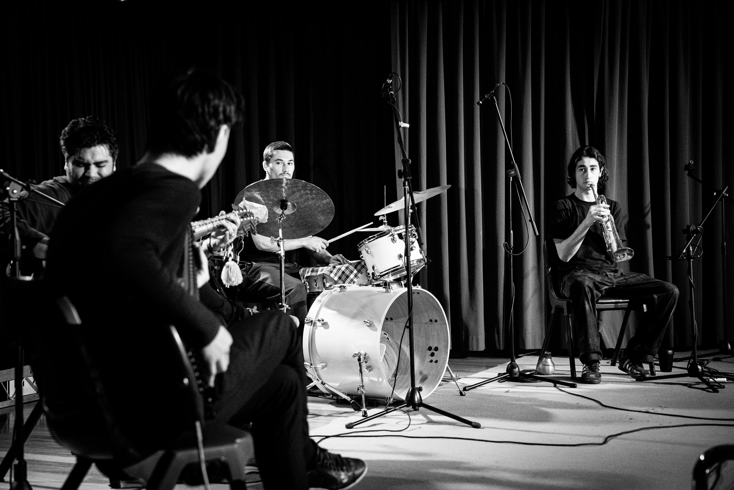 Black and white photo of a four-member jazz band performing on stage. The musicians include a drummer, a trumpet player, and two guitarists. They are surrounded by microphones and the background features stage curtains.