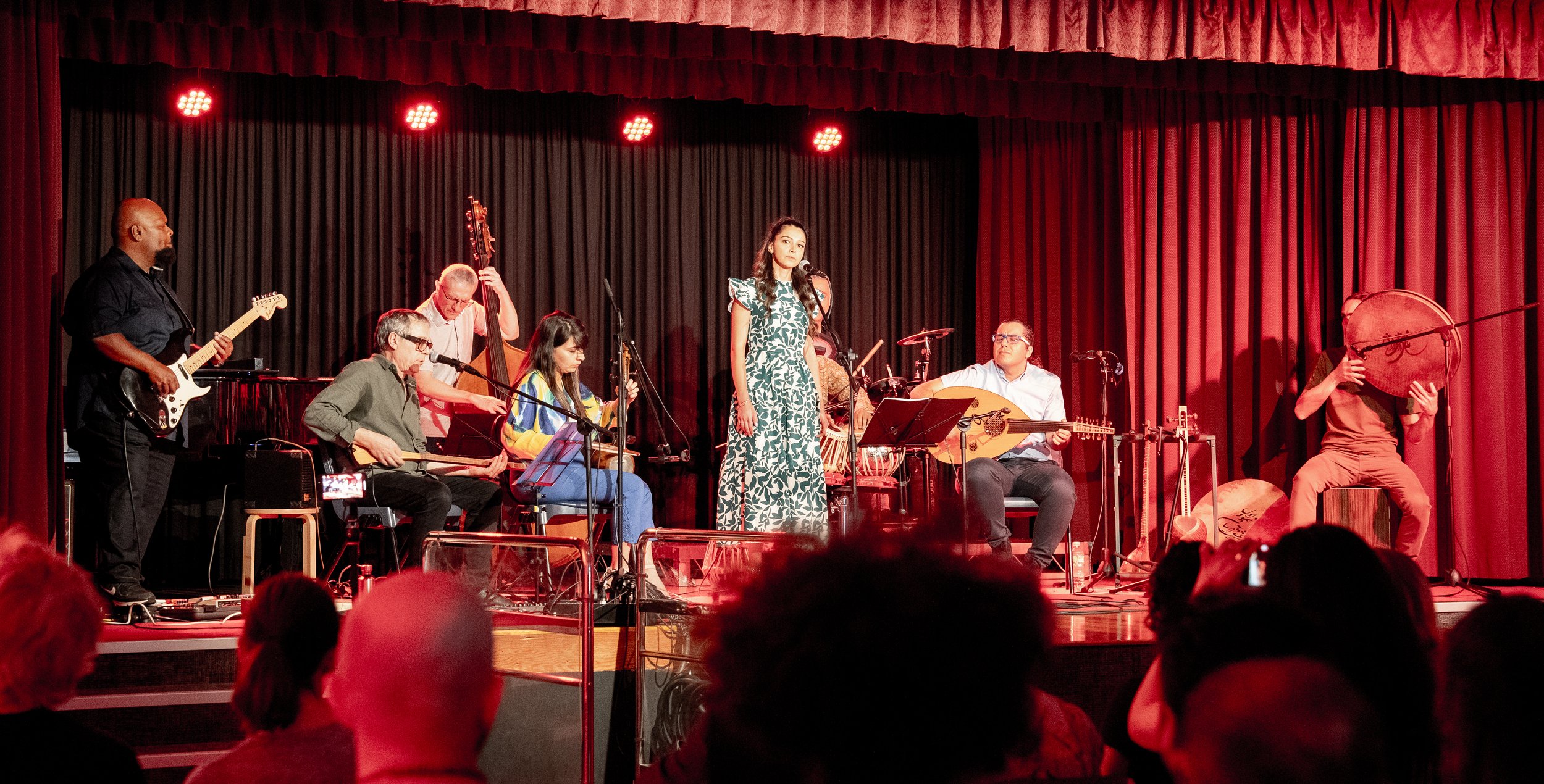 Band performing on stage with various instruments including guitar, oud, and drums, featuring a female singer, in front of a seated audience and red curtain backdrop.