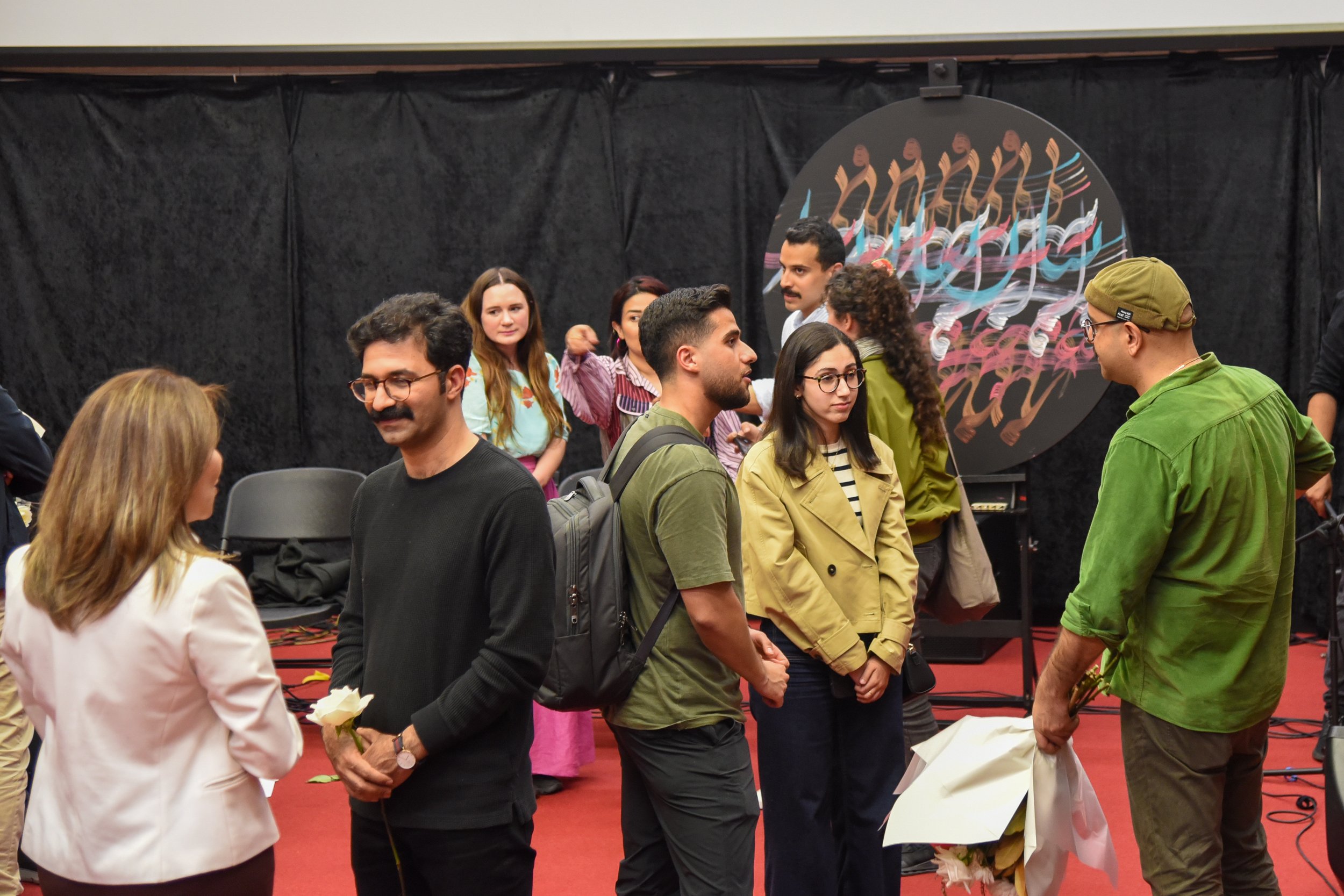 Group of people at an indoor event with artwork in the background, holding flowers and conversing.