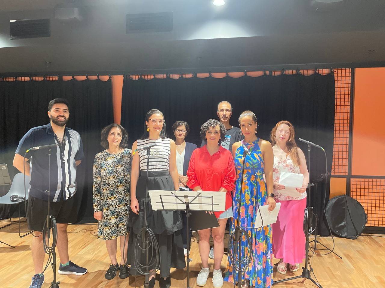 A group of eight people standing on a wooden floor in a room with microphones and music stands. They are dressed in casual and semi-formal attire, with colorful and patterned outfits. The background features black curtains and orange walls.