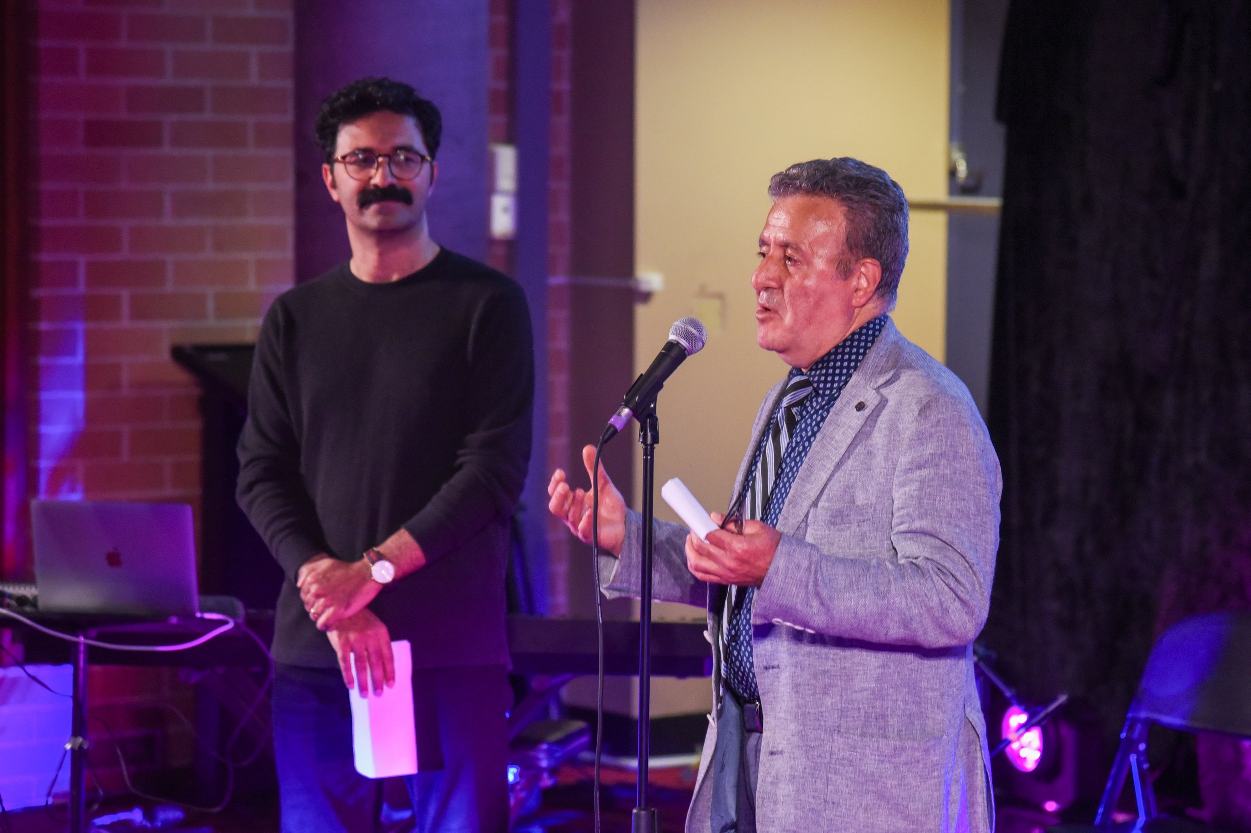 Two men in a room with a microphone and laptop, one speaking, one standing.