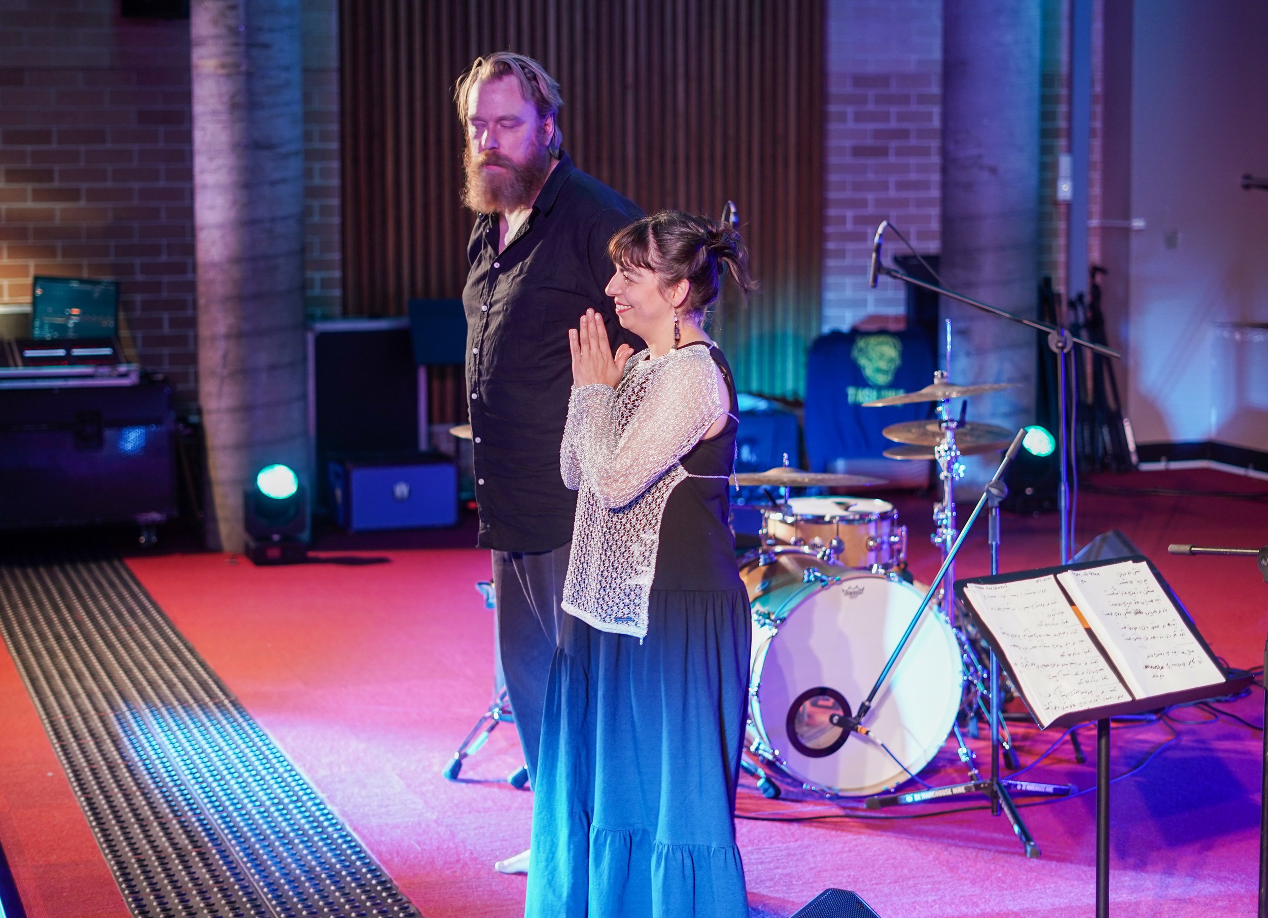 Two musicians standing on stage next to each other, with a drum set and sheet music in the background.