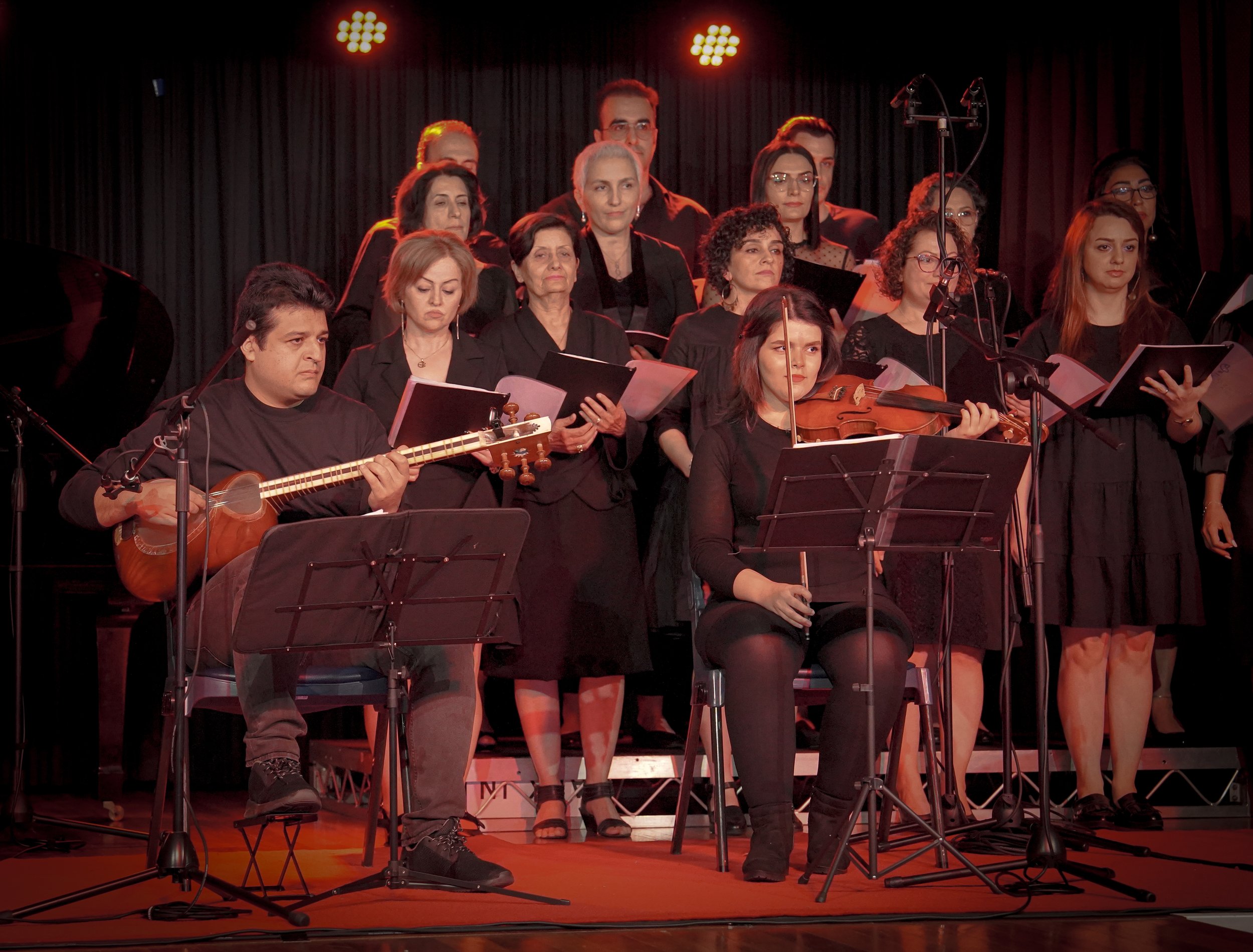 A choir performing on stage with two musicians in the foreground, one playing a string instrument and the other a violin, all dressed in black. Red stage lighting illuminates the scene.