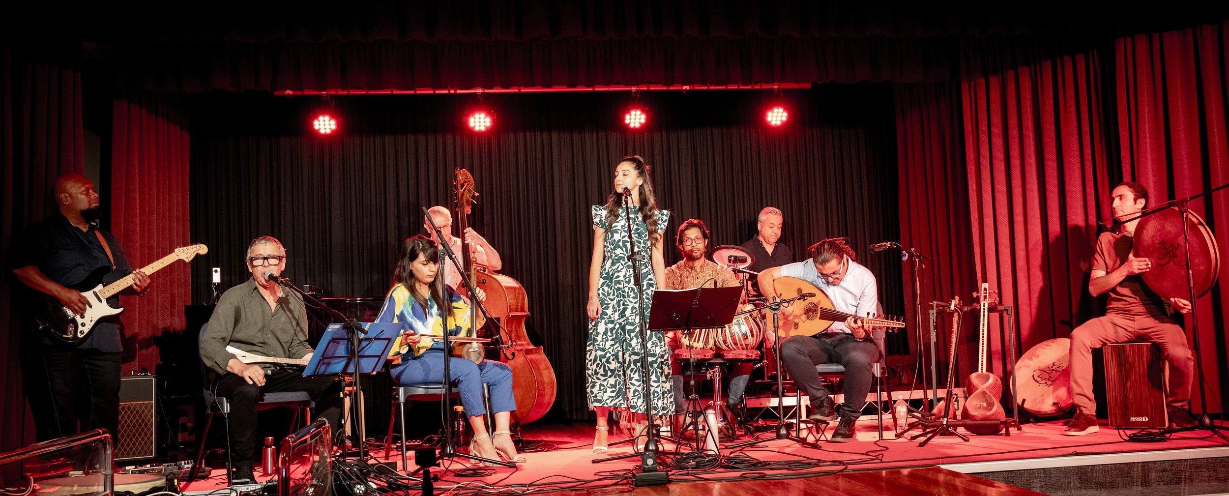 Live band performing on stage with a singer and various musicians playing instruments, including guitar, cello, percussion, and oud, under red stage lighting.