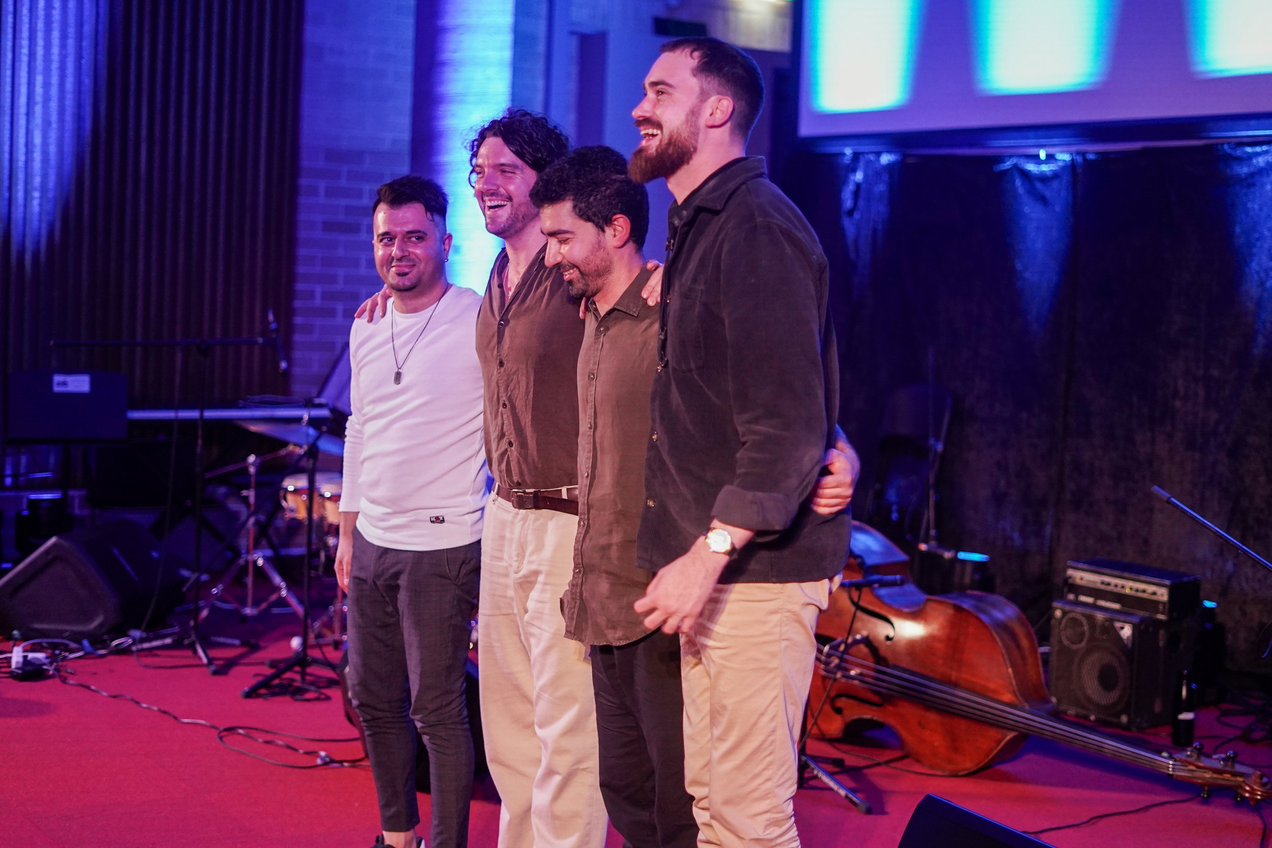 Four men standing together on a stage with instruments in the background, under blue lighting.