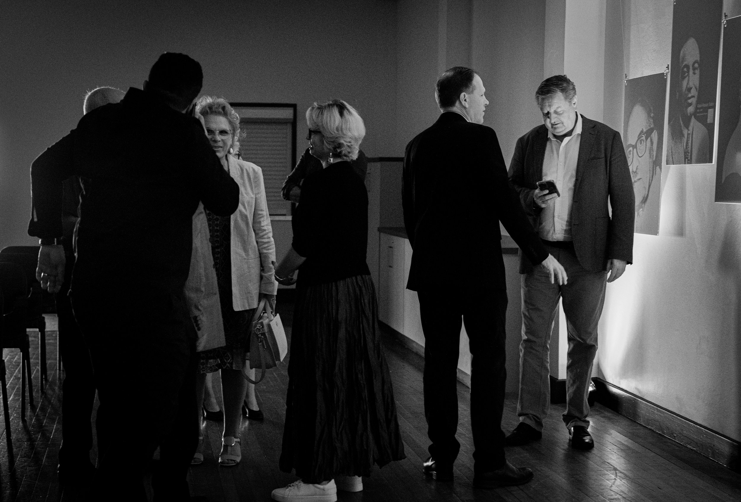 A black and white photo of a group of people in a room with photographs on the wall. One person is looking at a phone, while others are engaged in conversation. The setting appears formal, with individuals dressed in suits and dresses.