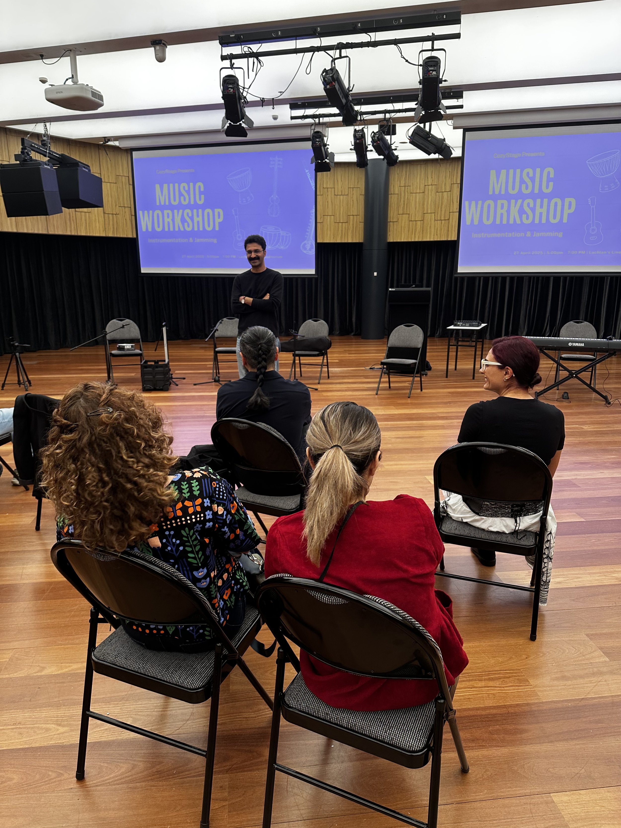 People sitting on chairs in a music workshop with a presenter standing in front of two screens displaying the text 'Music Workshop.'