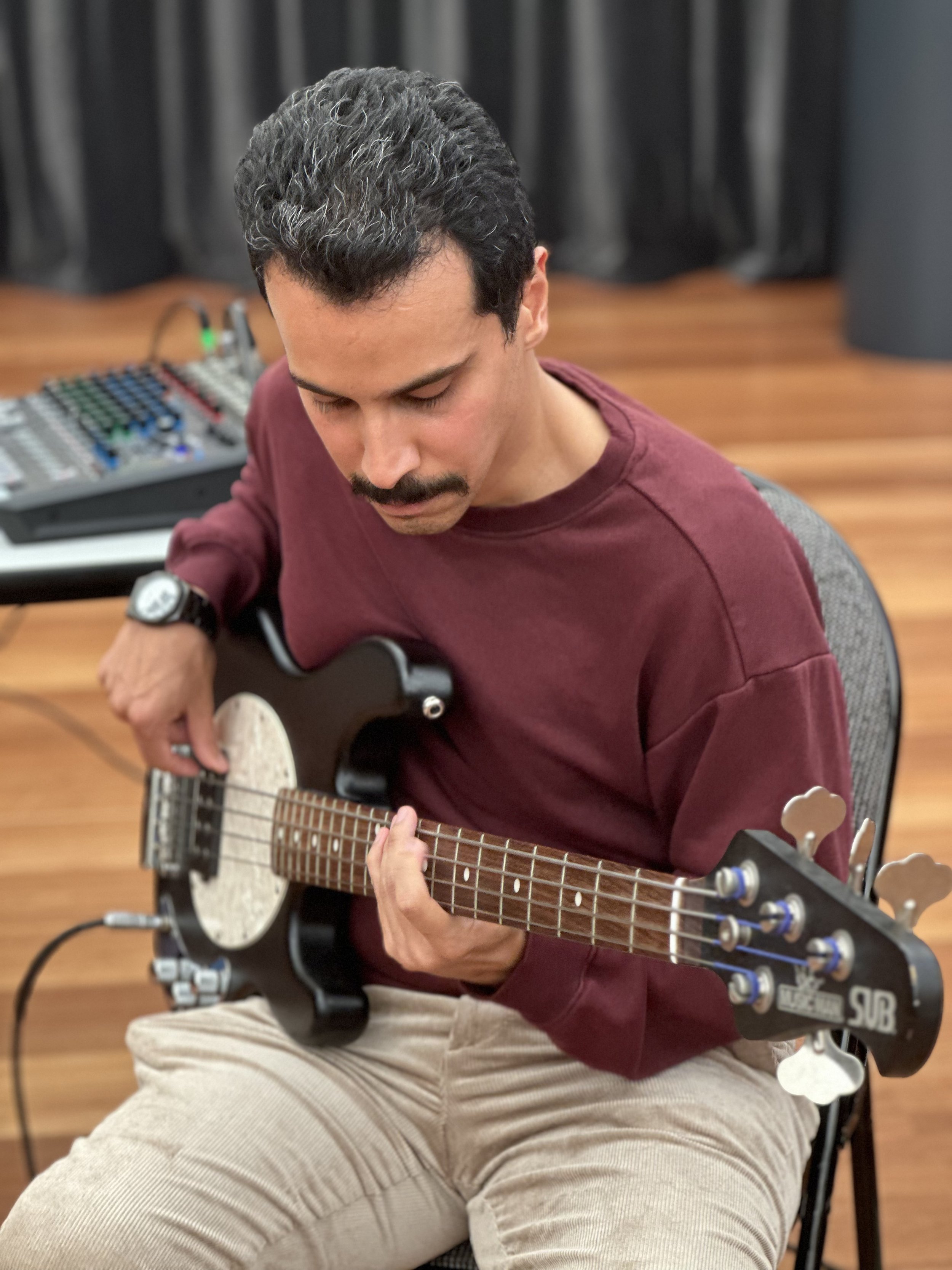 A person playing a black electric bass guitar, wearing a maroon sweater and beige pants. There is audio mixing equipment on a table in the background and a wooden floor beneath.