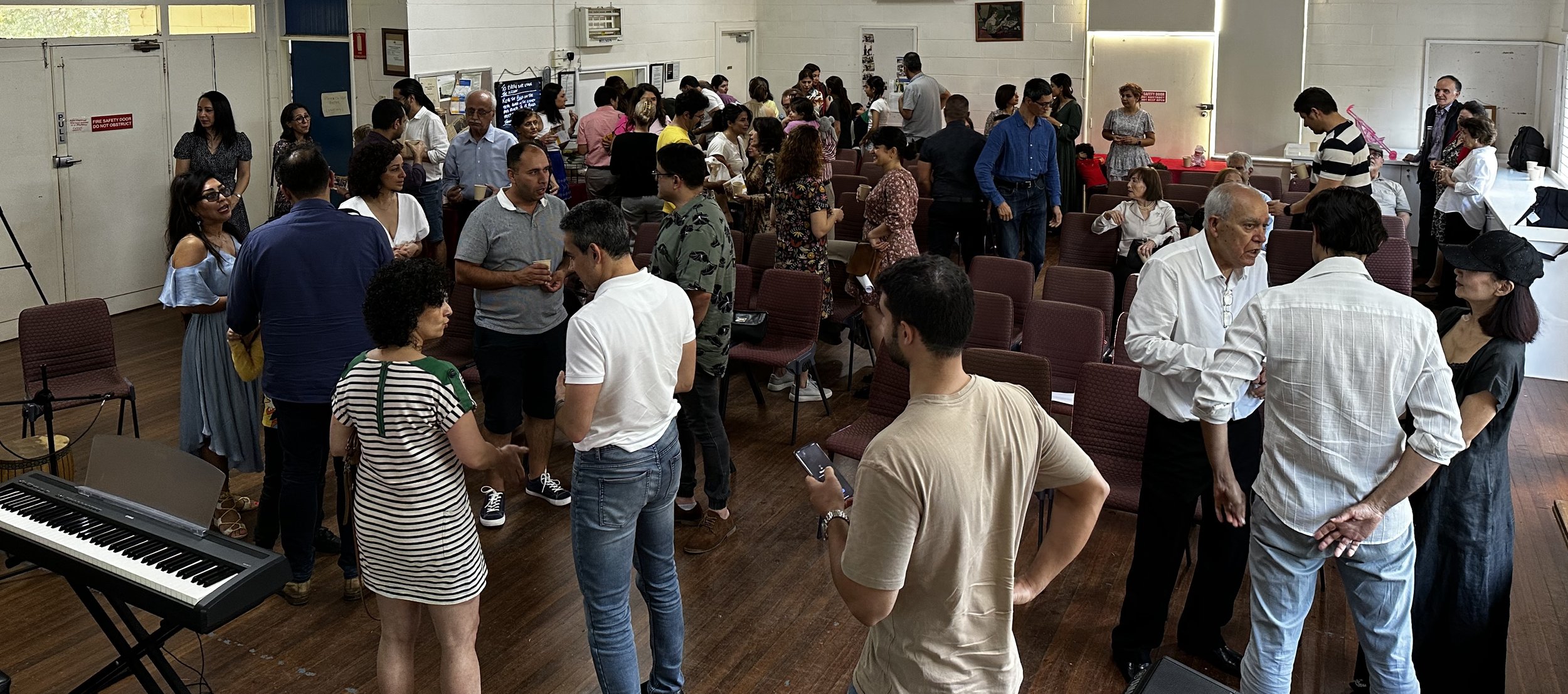 A group of people socializing in an indoor setting with chairs, a keyboard, and microphones visible.