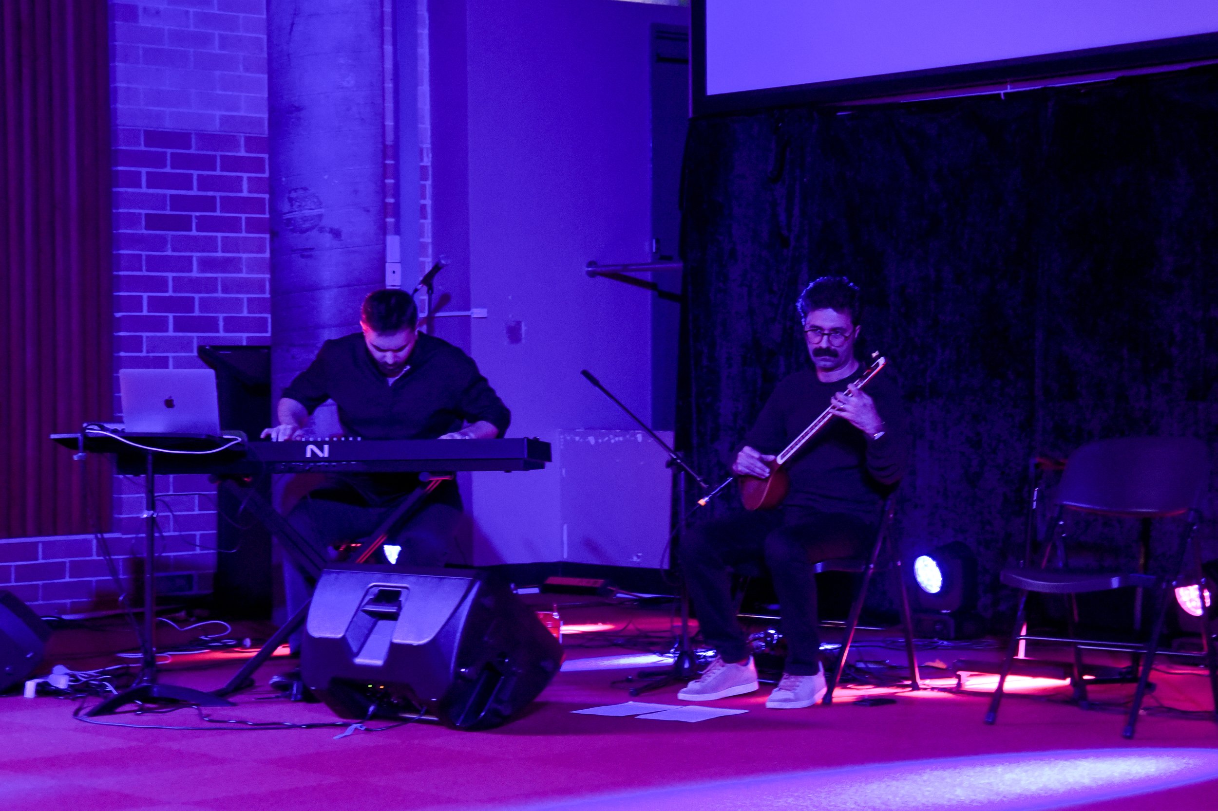 Two musicians performing on stage, one playing a keyboard and the other playing a string instrument. The stage is lit with purple and red lights.